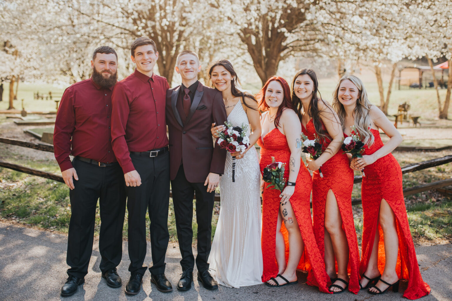 Elopement Wedding A group of six people stand outdoors in front of blossoming trees. Two men wear maroon shirts, and a man in the center wears a dark suit with a light-colored tie. A woman in a white dress stands beside him, flanked by four women in red dresses, all holding bouquets. They are smiling and posing, capturing the joy of an intimate elopement. Elopements Inc