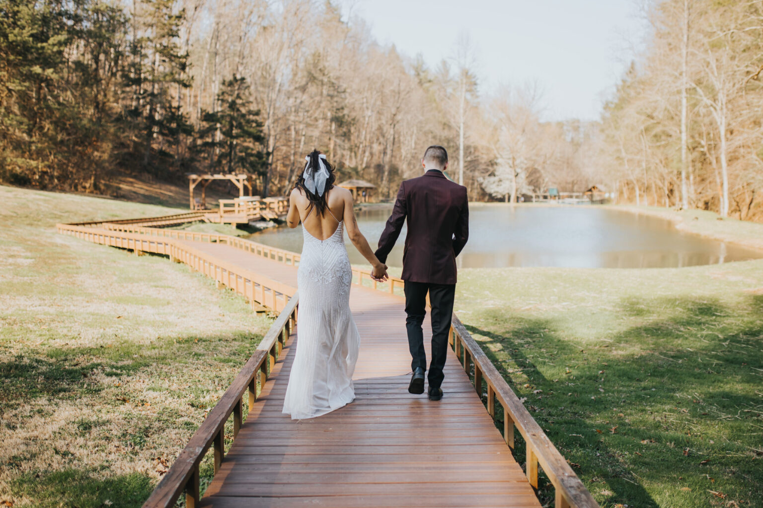 Elopement Wedding A couple walks hand-in-hand on a wooden pathway beside a serene pond. The woman in a white lace dress with a ribbon in her hair, and the man in a dark suit, are surrounded by lush grass and tall trees, suggesting an intimate elopement or special occasion in a beautiful outdoor setting. Elopements Inc