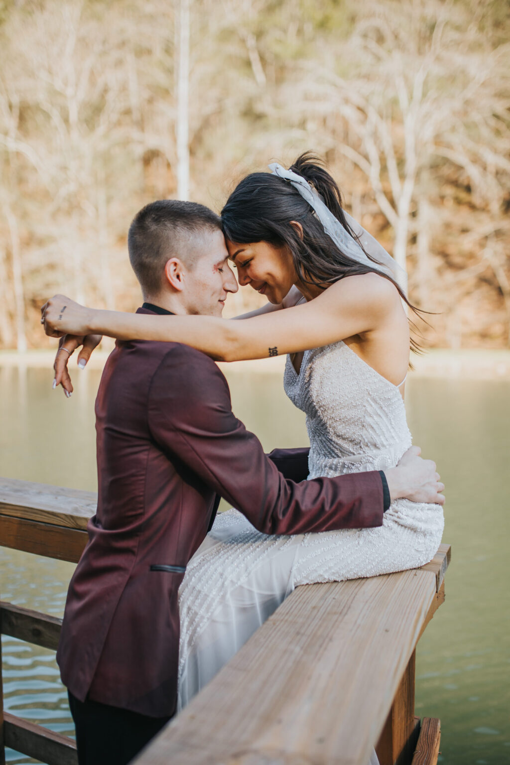 Elopement Wedding A newlywed couple shares an intimate moment on a wooden railing by a serene lake, having chosen to elope for their special day. The bride, in a white strapless dress and veil, embraces the groom, who is wearing a maroon suit. They press their foreheads together, eyes closed, surrounded by the natural setting of trees. Elopements Inc