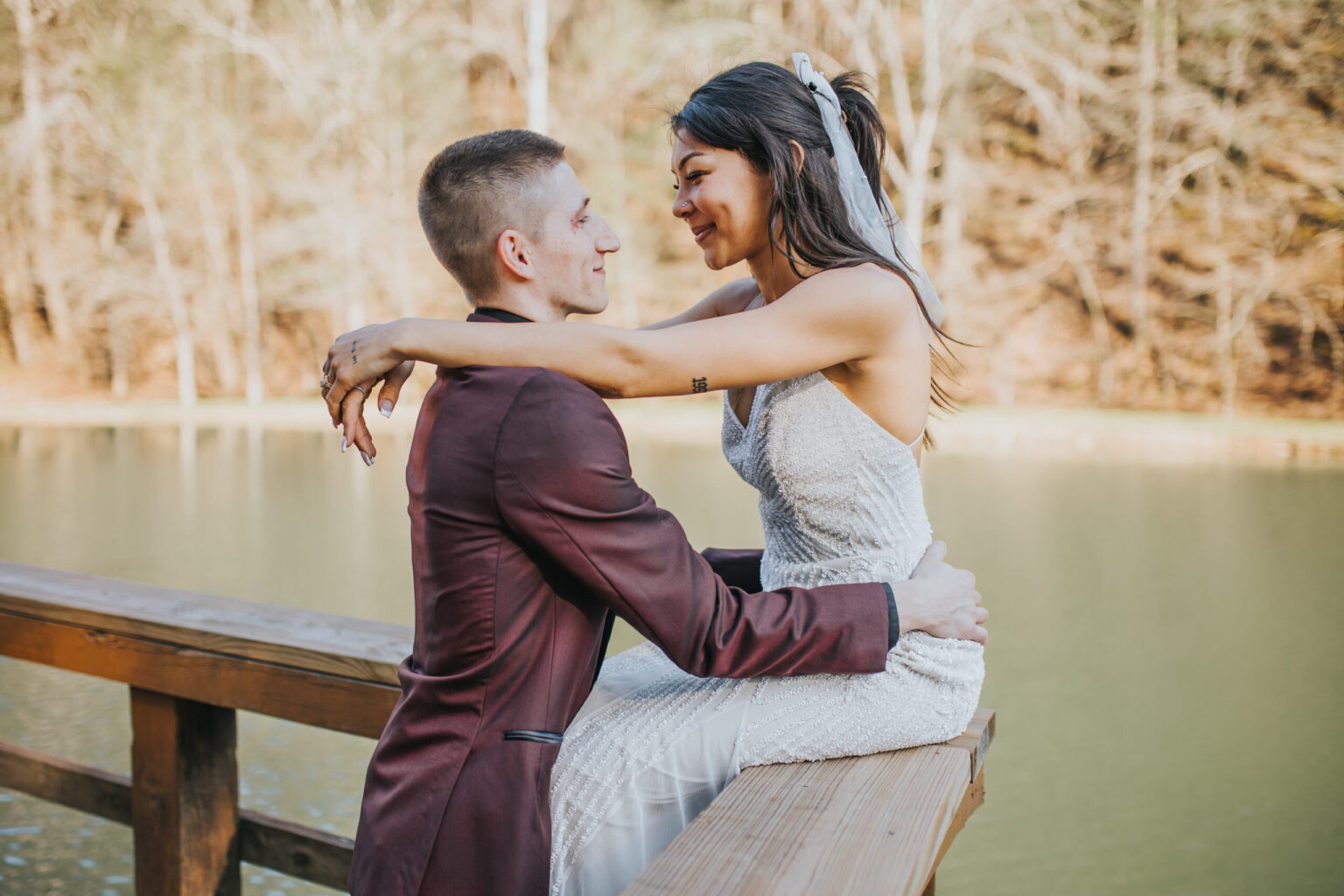 Elopement Wedding A newlywed couple sits close together on a wooden railing by a serene lake. The woman, in a white lace wedding dress with a veil, wraps her arms around the man's neck. The man, in a maroon suit, holds her waist. Both smile affectionately at each other amidst the autumn trees, perfect for an elopement setting. Elopements Inc