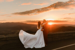 Elopement Wedding A bride in a flowing white gown and a groom in a dark suit embrace joyfully on a stone lookout, with a stunning sunset and mountains in the background. The sky is painted with warm hues of orange and pink, casting a romantic glow over their intimate elopement. Elopements Inc