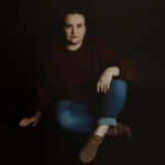 Elopement Wedding A woman with light skin and dark hair sits on the floor against a dark backdrop. She is wearing a maroon sweater, rolled-up blue jeans, and tan ankle boots. She has a neutral expression, one leg crossed over the other, with her right hand resting on her knee and her left hand on the floor. Elopements Inc