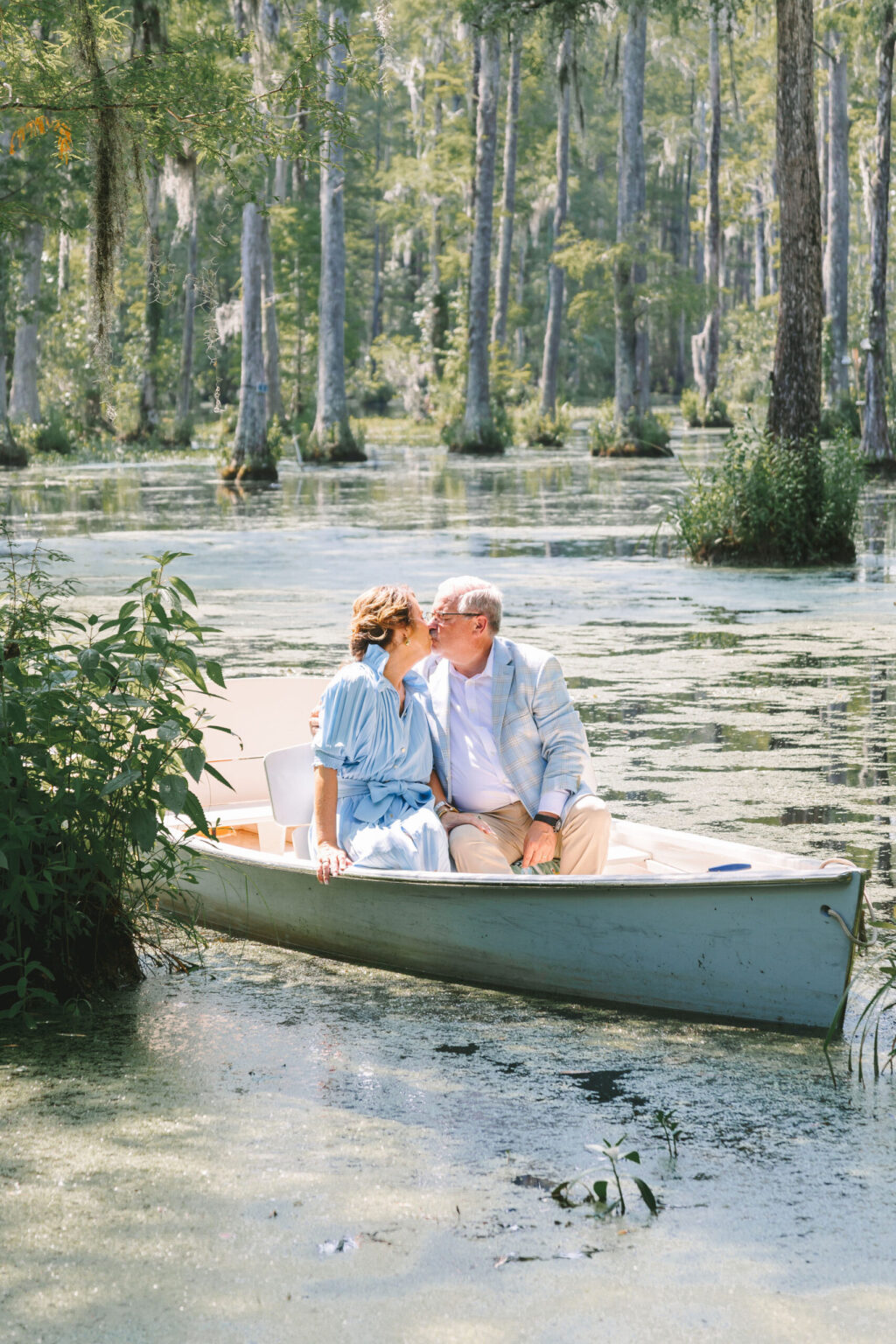 Elopement Wedding A couple sits in a rowboat on a calm, forested lake. They're dressed in light clothing and share a kiss, surrounded by green trees and water covered in patches of algae. The serene setting of cypress trees and hanging moss makes their elopement feel deeply romantic and tranquil. Elopements Inc