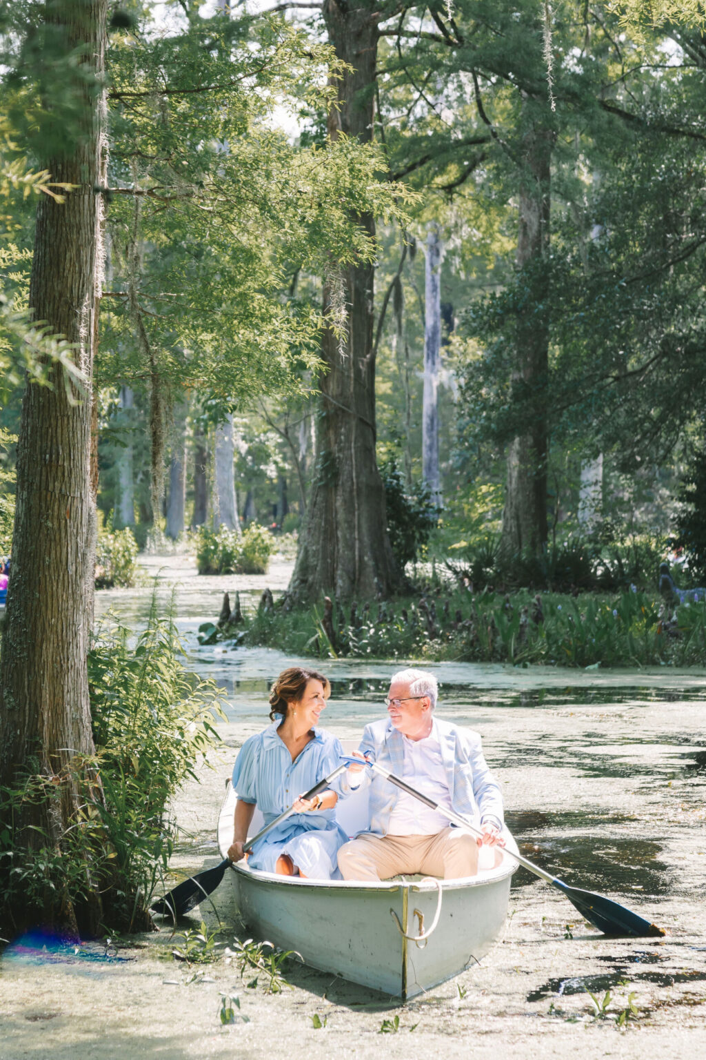 Elopement Wedding A woman and an older man sit in a small rowboat on a serene, tree-lined lake. The woman, in a light blue dress, paddles with an oar, while the man, wearing a light blue jacket and glasses, holds another oar. They both look at each other smiling as sunlight filters through the trees—an elopement to remember. Elopements Inc