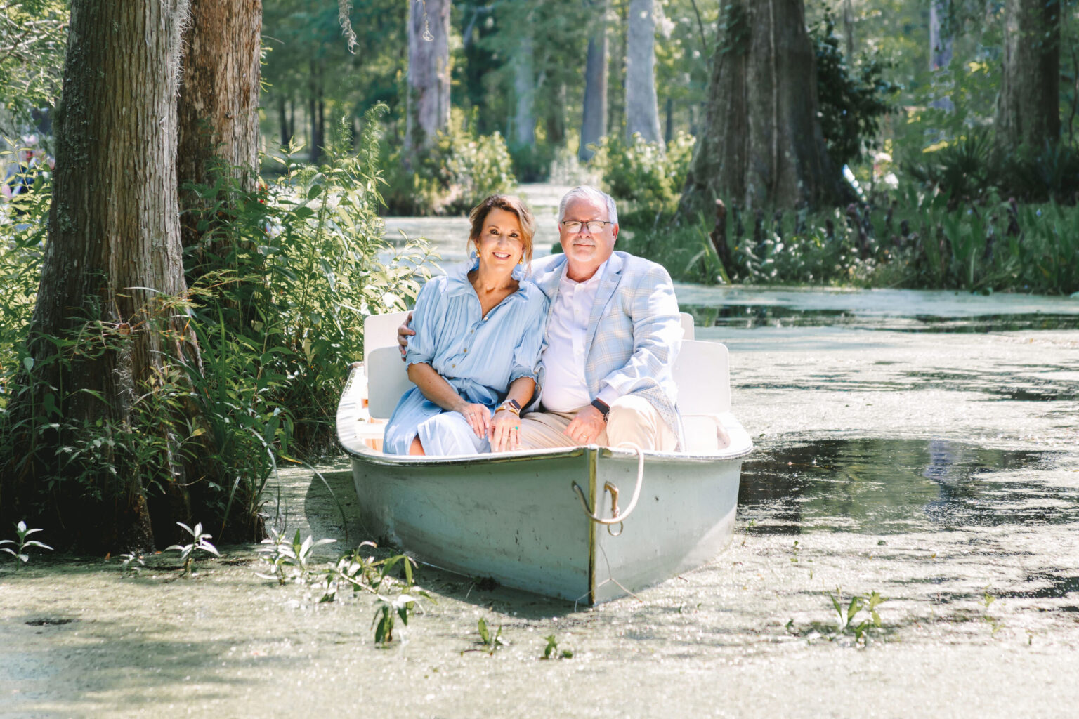 Elopement Wedding A woman and a man sit side by side in a small, white boat on a calm, greenish river surrounded by lush trees and vegetation. They are both smiling and appear to be enjoying a relaxing elopement outdoors. The woman is wearing a light blue dress, while the man is dressed in a light beige suit. Elopements Inc