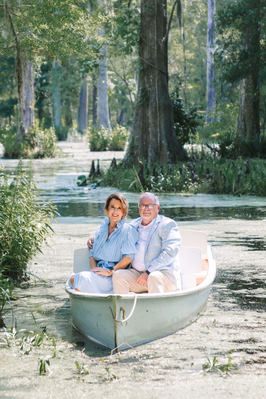 Elopement Wedding A man and woman who recently decided to elope sit in a small rowboat on a tranquil, tree-lined lake. Both are smiling and dressed in light-colored, casual clothing. The lake is surrounded by lush greenery and tall trees, with sunlight filtering through the branches, creating a serene and picturesque setting. Elopements Inc