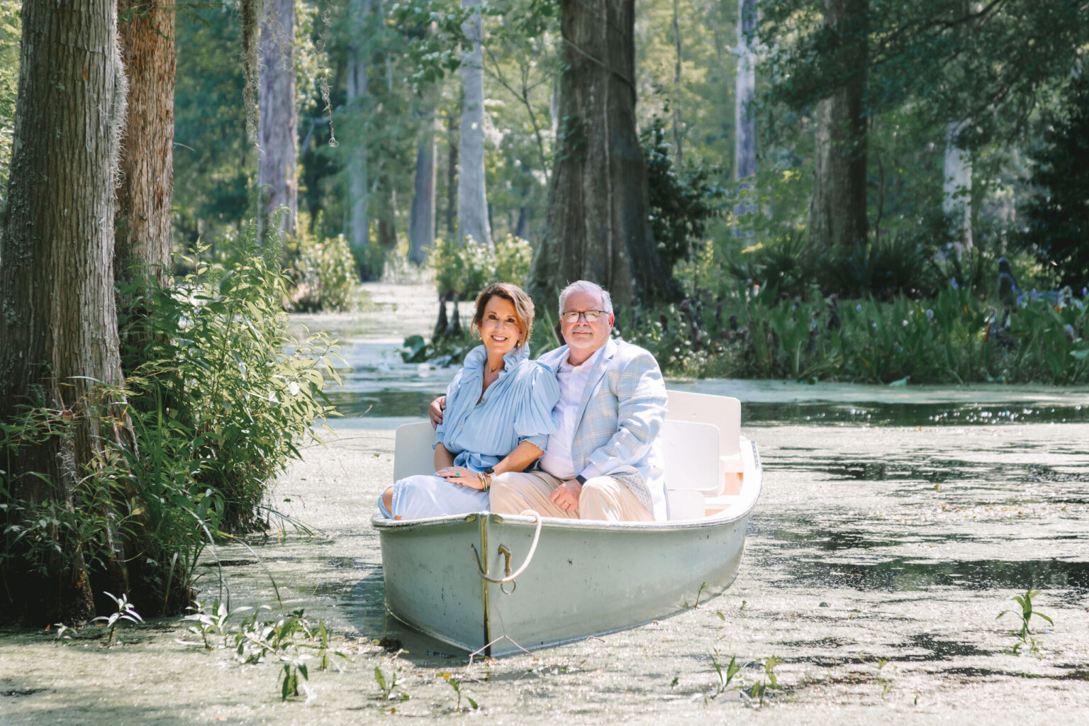 Elopement Wedding A couple enjoys a serene moment on a light blue rowboat, eloping amidst lush greenery and trees in a peaceful, narrow waterway. The woman is wearing a light blue dress, while the man dons light-colored pants and a plaid blazer. Sunlight filters through the trees, illuminating their secretive elopement. Elopements Inc