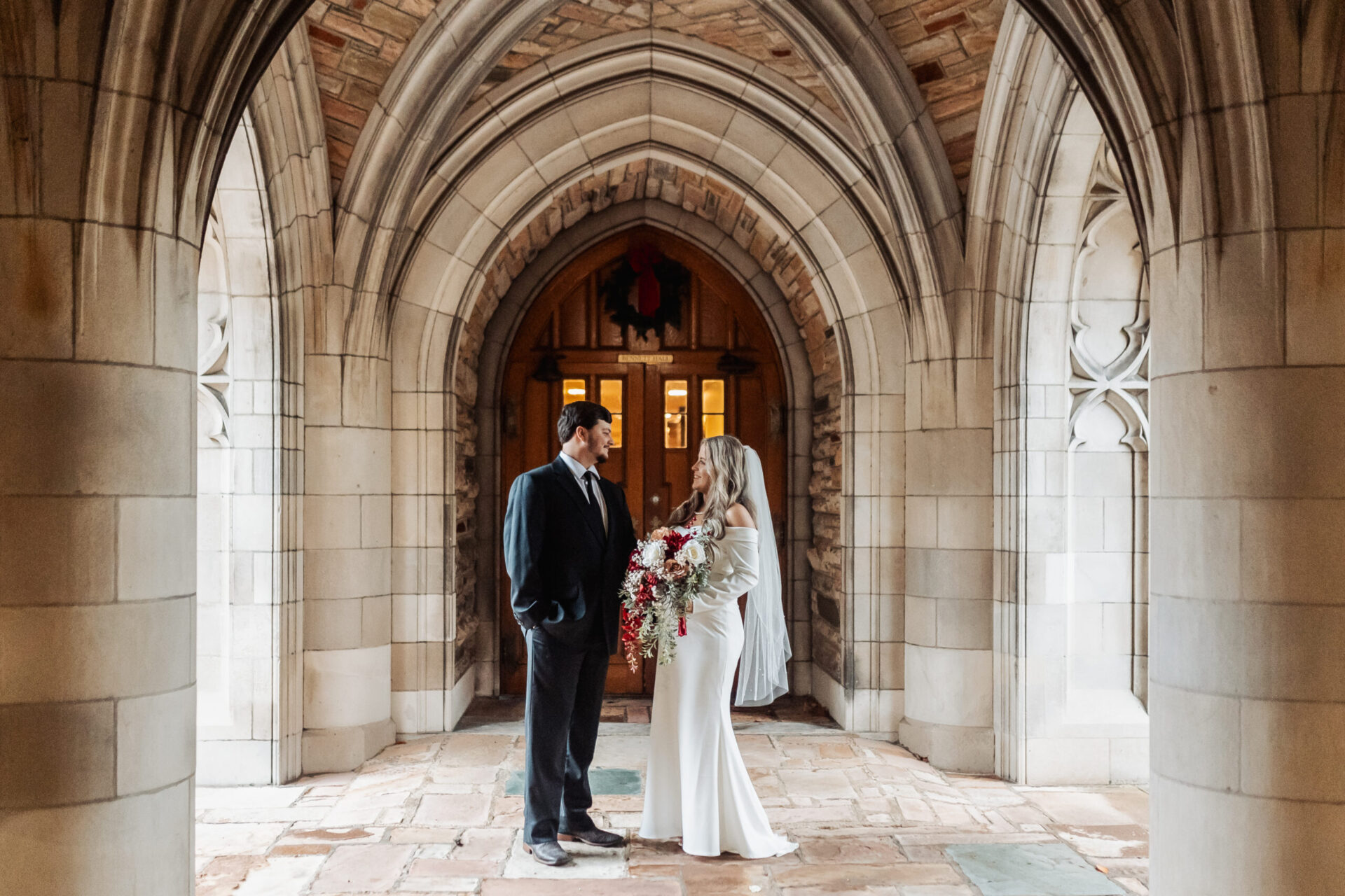 Elopement Wedding A bride and groom stand facing each other under a stone archway of an ornate building. The bride, in a white dress and veil, holds a large bouquet of flowers. The groom wears a dark suit. The arched doorway behind them is intricately designed, creating a beautiful, timeless backdrop for their elopement. Elopements Inc