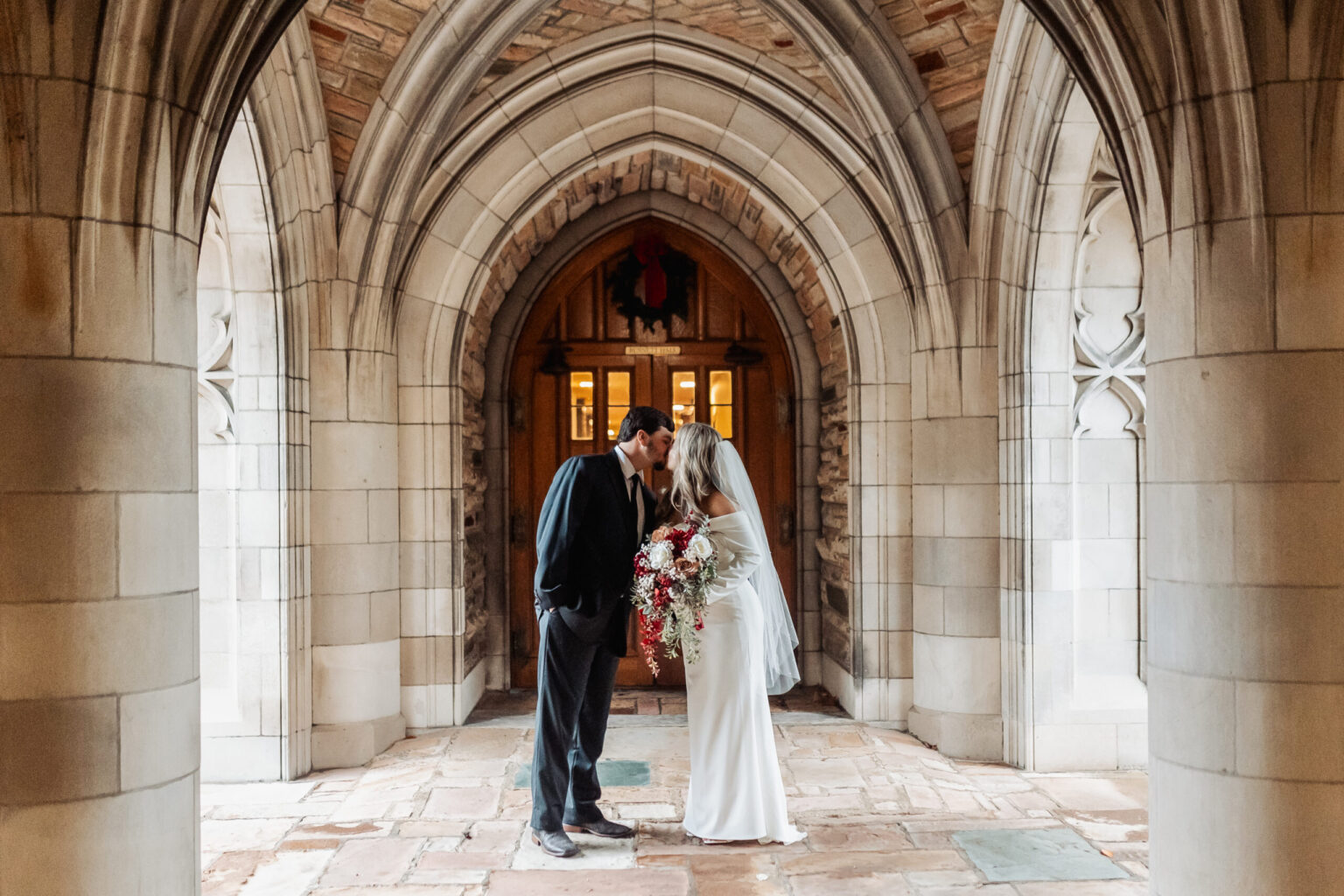 Elopement Wedding A bride and groom share a kiss beneath a stone archway, surrounded by gothic-style architecture. The bride holds a large bouquet with white and red flowers and is dressed in a long white gown with a veil. The groom, in a dark suit, leans in, touching his head to hers—a moment of intimate elopement perfection. Elopements Inc