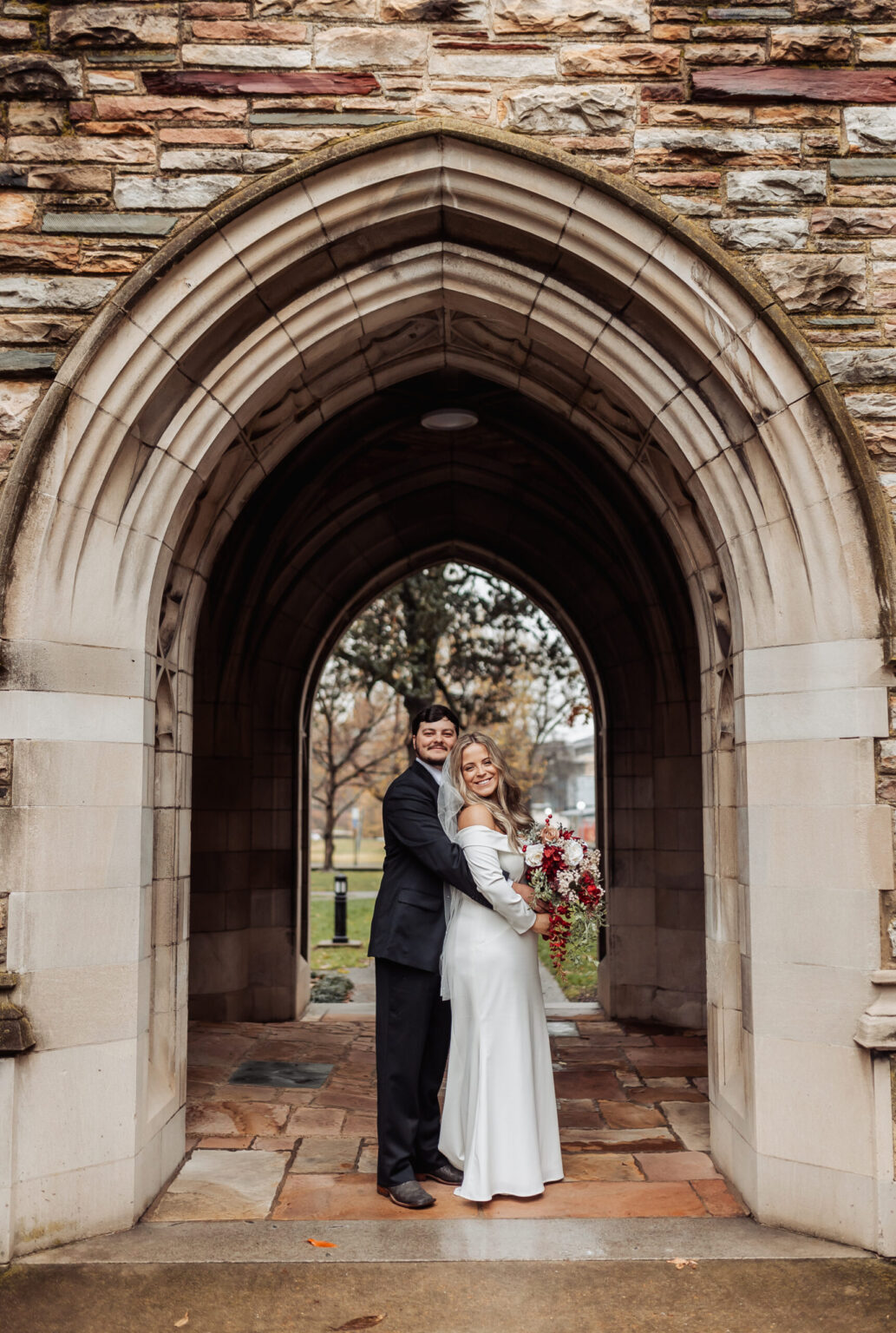 Elopement Wedding A newly married couple stands under a stone archway. The groom, in a black suit, hugs the bride from behind. She wears a long white dress and holds a bouquet of red, white, and green flowers. The couple smiles joyfully. Their intimate elopement perfectly complements the rustic, historic feel of the archway's stone structure. Elopements Inc