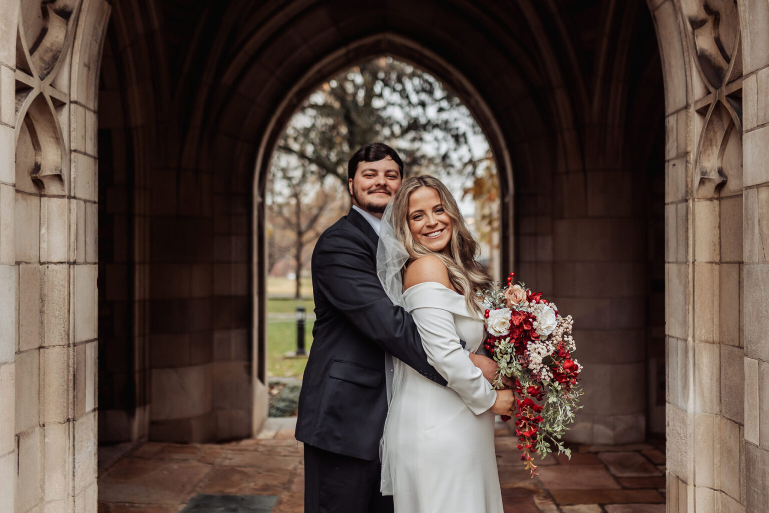 Elopement Wedding A smiling couple is posing for a photo under a stone archway. The man in a dark suit is standing behind the woman in an off-the-shoulder white dress, embracing her. She is holding a vibrant bouquet with red, white, and pink flowers. The background features greenery and a tree, perfect for an elopement. Elopements Inc