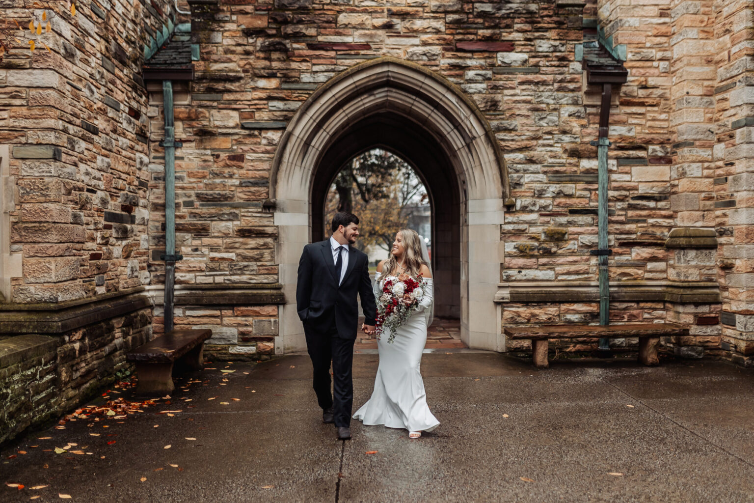 Elopement Wedding A couple, possibly having chosen to elope, walks hand in hand beneath a stone archway. The man is in a black suit and tie, while the woman wears a white gown and holds a bouquet of red and white flowers. They smile at each other against the backdrop of a stone building with Gothic architecture. Elopements Inc