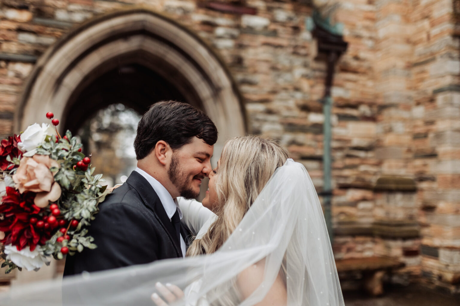 Elopement Wedding A bride and groom, possibly eloping, stand close together in front of a stone archway. The bride, wearing a veil, holds a bouquet of red and white flowers. The groom, dressed in a dark suit, gazes affectionately at her as they touch foreheads, smiling warmly. The scene is romantic and intimate. Elopements Inc