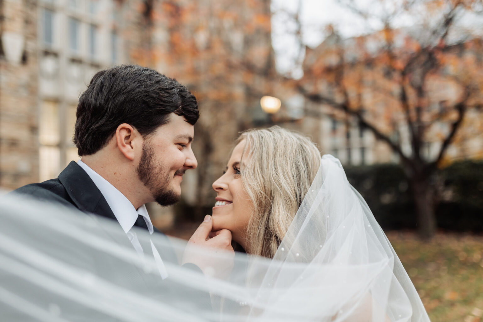 Elopement Wedding A bride and groom gaze lovingly at each other outdoors during their intimate elopement. The groom, wearing a black suit, gently holds the bride's chin. The bride, in a white dress with a veil, smiles up at him. Autumn leaves and a building provide a blurred backdrop, creating a romantic atmosphere. Elopements Inc