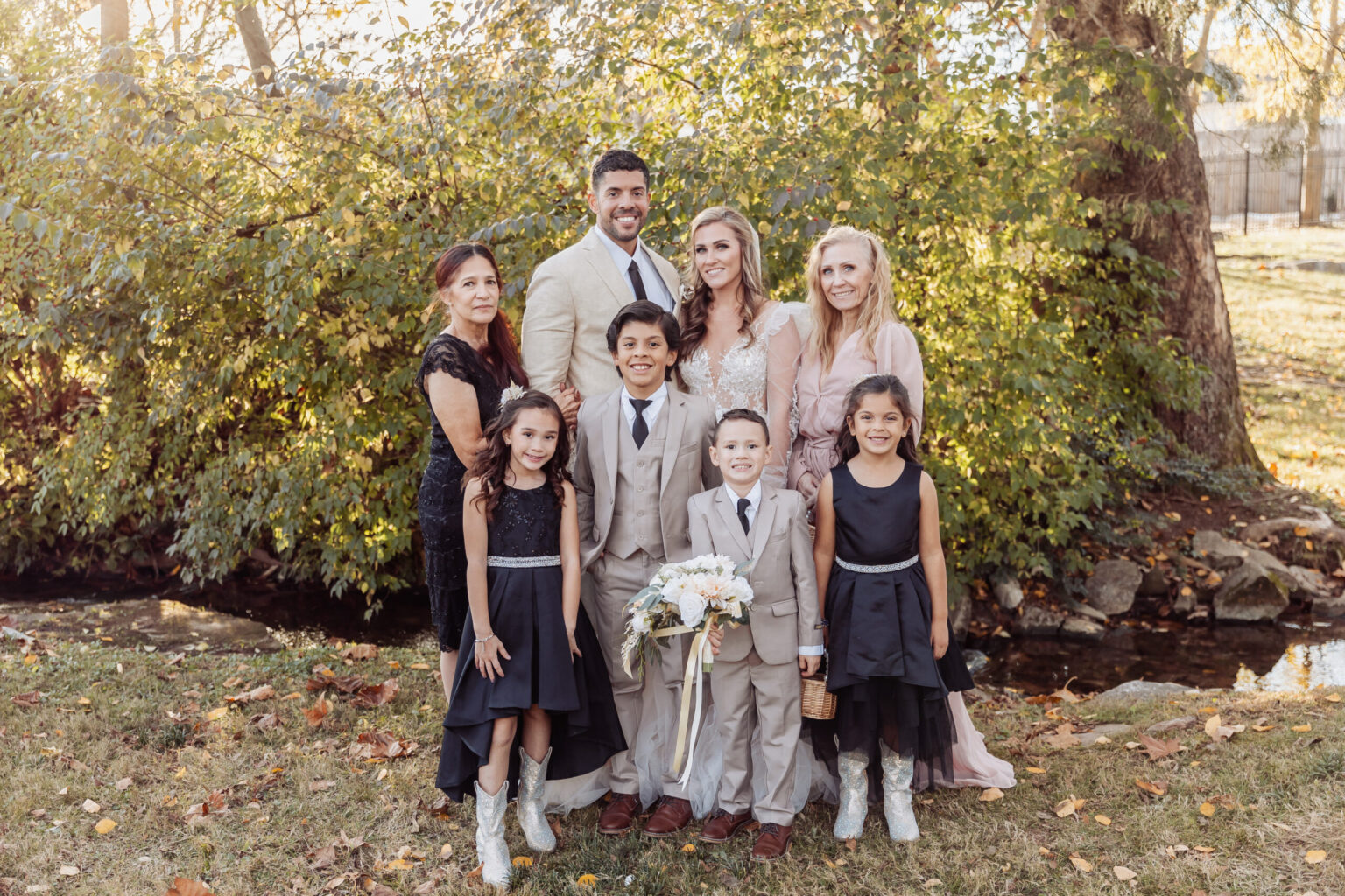 Elopement Wedding A joyful elopement portrait features a smiling couple with five children and two older women, posing outdoors near a creek with leafy bushes as the backdrop. The children are dressed in black dresses and tan suits, with the bride in a white gown and the groom in a beige suit. Elopements Inc