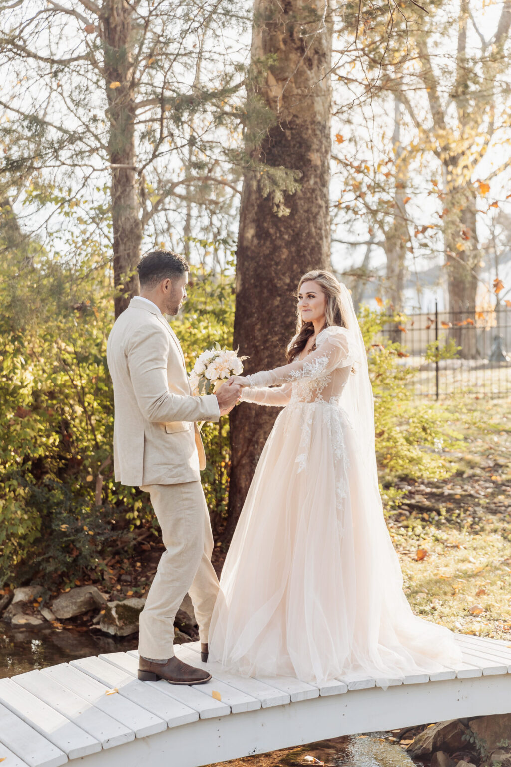 Elopement Wedding A bride and groom stand on a small white wooden bridge, surrounded by greenery and trees. The bride wears a long, white dress with lace details and a veil, holding a bouquet. The groom is in a light beige suit. They hold hands, looking at each other with the warmth of elopement in the air. Elopements Inc