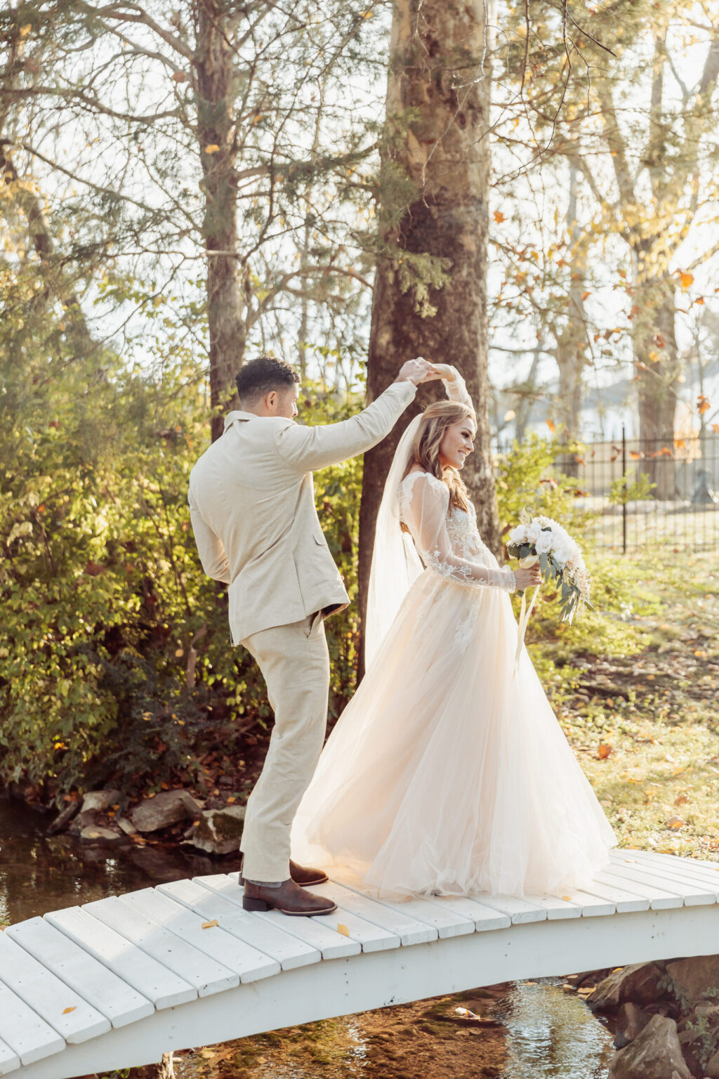 Elopement Wedding A groom in a light suit twirls his bride, who is in a flowing white wedding gown, on a small white wooden bridge. The couple eloped and is now surrounded by autumn trees with golden leaves and soft sunlight filtering through. The bride holds a bouquet of white flowers, and a small stream runs beneath the bridge. Elopements Inc