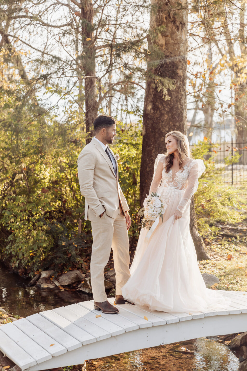 Elopement Wedding A bride and groom, who decided to elope, stand on a small white wooden bridge in a serene, wooded setting. The bride is holding a bouquet, wearing an elegant, flowing white dress with lace details and puffed sleeves. The groom is dressed in a beige suit with a black tie. Rays of sunlight filter through the trees. Elopements Inc