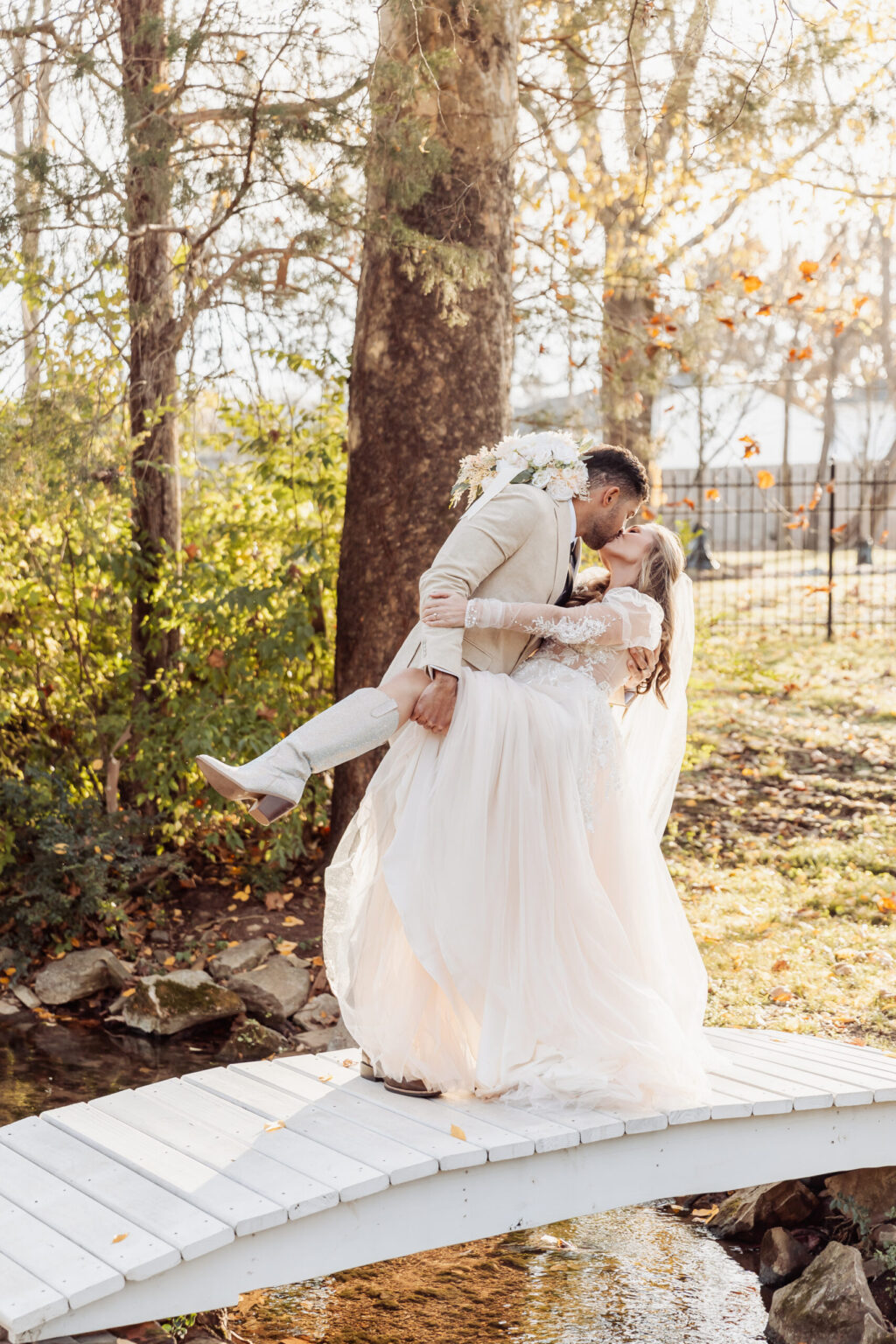 Elopement Wedding A groom in a light-colored suit lifts and kisses a bride in a flowing white gown and veil on a small white footbridge over a stream. Trees surround the eloping couple with autumn foliage in the background, creating a romantic outdoor setting. Elopements Inc