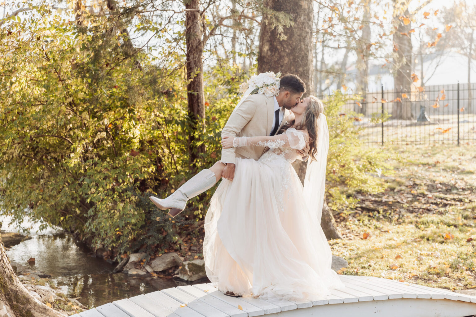 Elopement Wedding A groom in a tan suit dips and kisses his bride in a white lace wedding dress and a long veil on a small wooden bridge in a sunlit forest. She holds a bouquet of white flowers, and he lifts her partially off her feet. Fall foliage and trees surround the couple, adding a warm, golden hue to their elopement. Elopements Inc