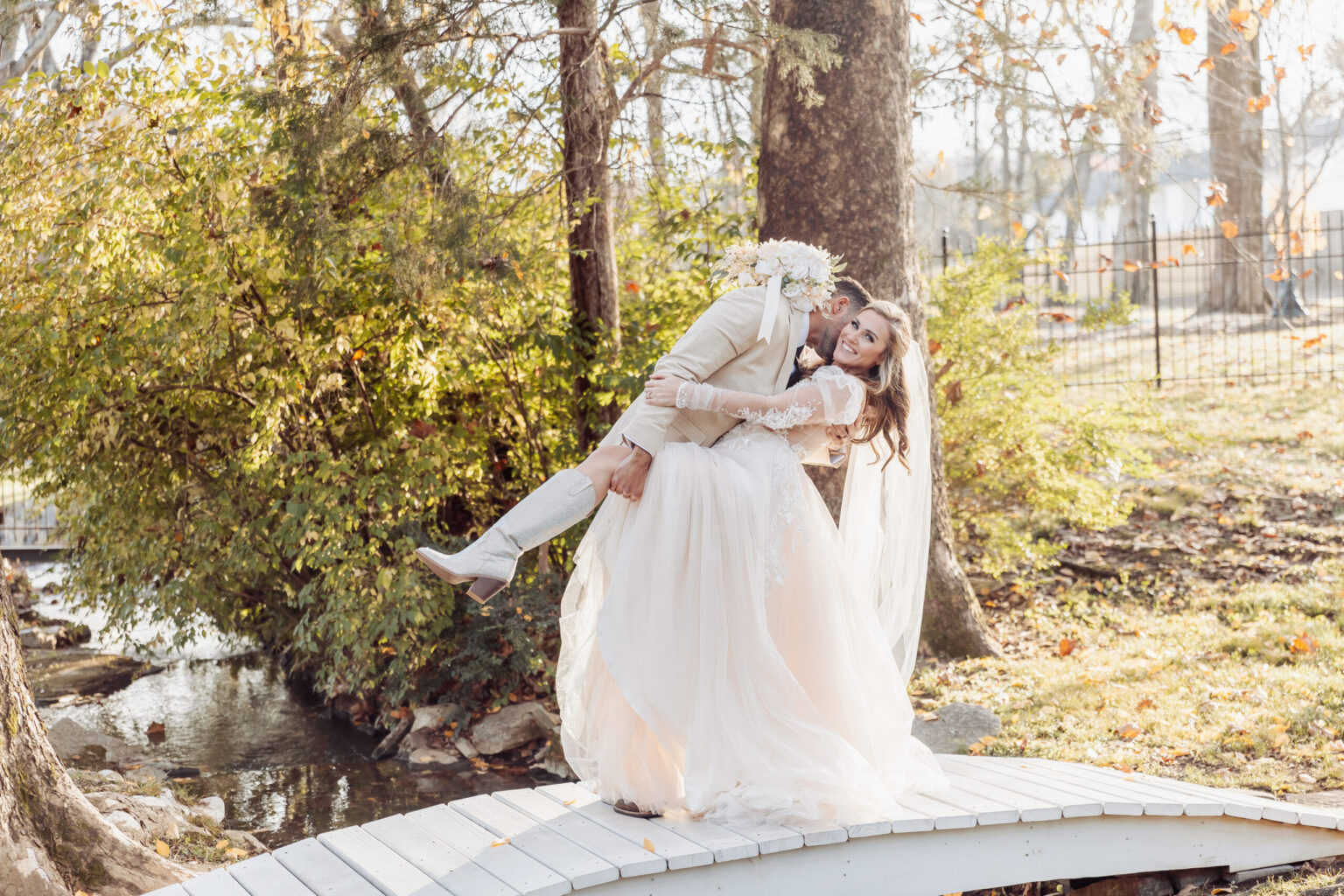 Elopement Wedding A bride and groom, who decided to elope, are standing on a small, arched wooden bridge in a park. The groom, in a light beige suit, playfully dips the bride in her white long-sleeved wedding gown with a veil. They are both smiling amidst green trees and sunlight filtering through. Elopements Inc