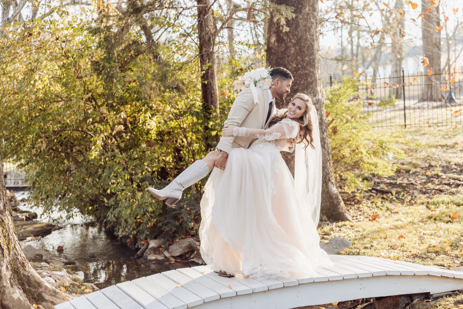 Elopement Wedding A groom, eloping with his bride, lifts her on a small white bridge in a garden. He wears a beige suit and bow tie, while she dons a long, flowing white dress with lace sleeves and a veil. Sunlight filters through the trees, highlighting the couple and the vibrant autumn foliage around them. Elopements Inc