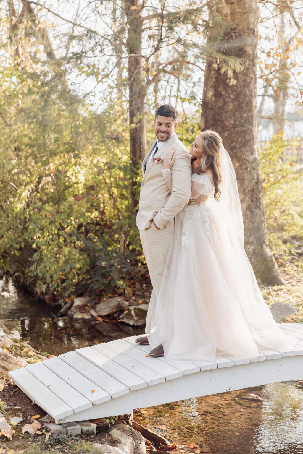 Elopement Wedding A bride and groom stand closely together on a small white wooden bridge over a stream in a wooded area. The groom, wearing a light beige suit, smiles and looks over his shoulder, while the bride, in a flowing white dress and veil, embraces him from behind, also smiling. Sunlight filters through the trees during their intimate elopement. Elopements Inc