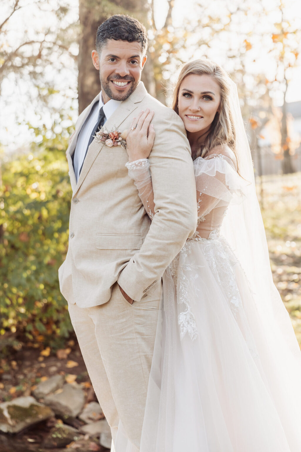 Elopement Wedding A smiling couple poses outdoors on their elopement day. The groom wears a beige suit with a white shirt and light blue tie, while the bride stuns in a white lace wedding gown with sheer sleeves and a veil. She stands behind him, leaning on his shoulder, amidst trees adorned with autumn foliage. Elopements Inc