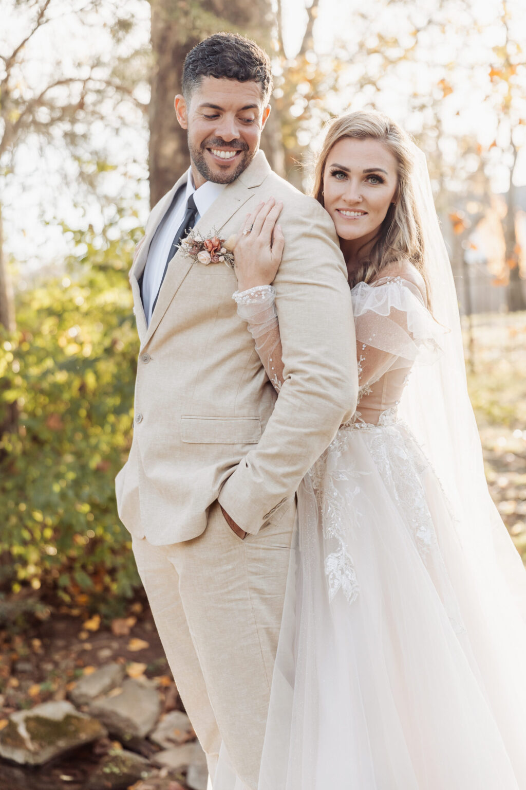 Elopement Wedding A smiling groom in a light beige suit with a boutonniere stands before his bride. The bride, in a lace-embellished wedding gown and veil, hugs him from behind while also smiling. They are outdoors with autumn foliage in the background, capturing the intimate joy of their elopement. Elopements Inc