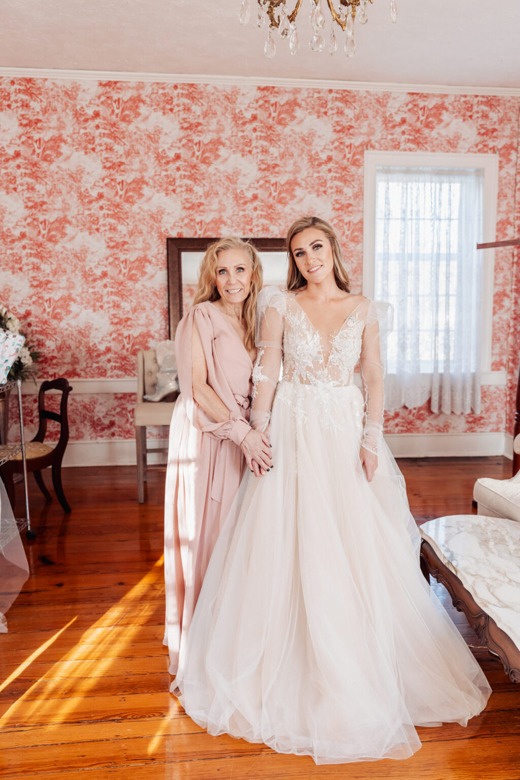 Elopement Wedding A bride and an older woman, likely her mother, stand smiling in a room with red and white floral-patterned wallpaper. The bride, perhaps planning an elopement, wears a white, long-sleeved wedding gown, while the older woman is dressed in a long, light pink dress. Sunlight streams in from a window, illuminating the hardwood floor. Elopements Inc