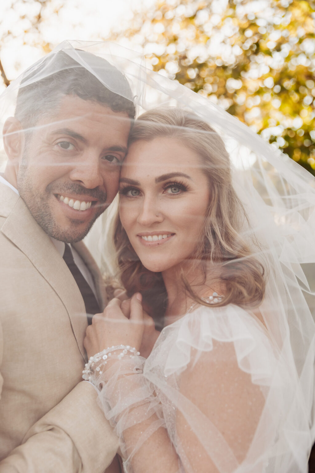 Elopement Wedding A bride and groom smile joyfully under a sheer, white veil. The groom, in a beige suit and white shirt, embraces the bride who has wavy blonde hair and wears a white dress adorned with small pearls. The background features soft, blurred greenery and sunlight filtering through the trees, capturing their intimate elopement. Elopements Inc