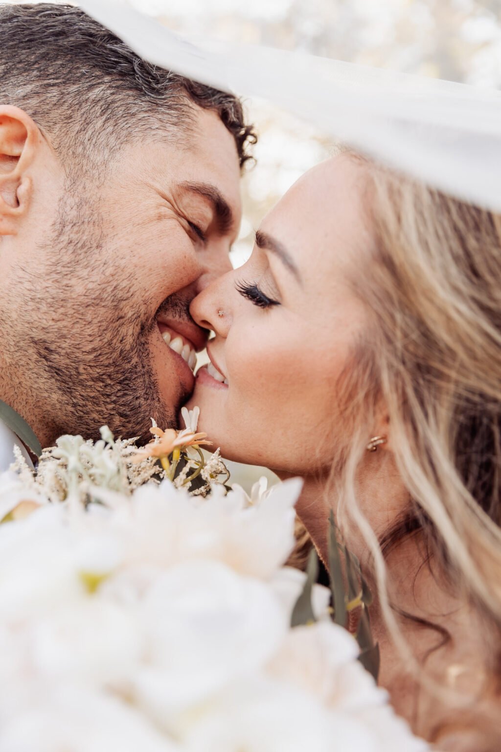 Elopement Wedding A close-up photo of a couple on their wedding day, affectionately touching noses and smiling with eyes closed. The groom has short, dark curly hair and a beard, while the bride has long, wavy blonde hair. Surrounded by soft, white flowers and a sheer veil draping over them, they embrace the intimacy of eloping. Elopements Inc