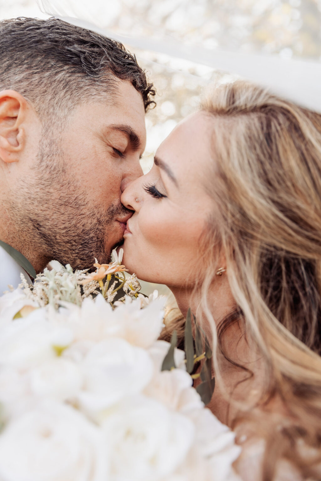 Elopement Wedding A close-up of a couple kissing tenderly during their elopement. The man on the left has short, curly hair and facial hair, while the woman on the right has long, wavy blonde hair. They hold a bouquet of light-colored flowers, mainly white and blush pink, capturing a romantic and intimate moment. Elopements Inc