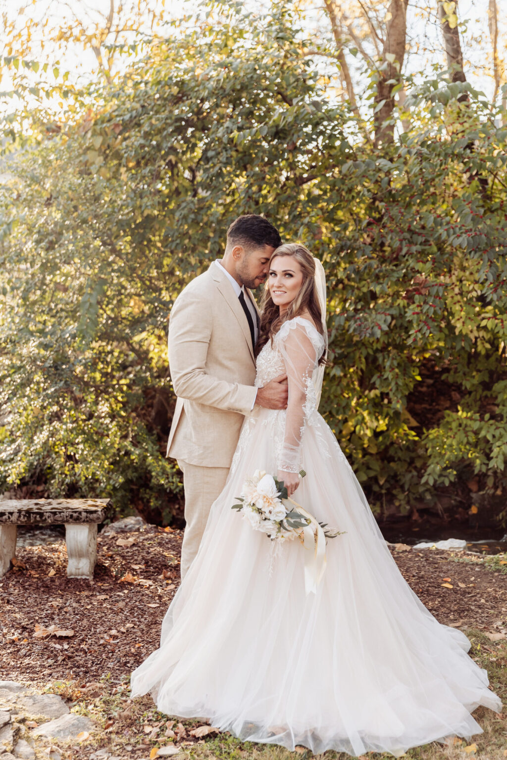 Elopement Wedding A couple poses for their elopement photo outdoors in front of greenery. The groom is wearing a beige suit with a white shirt, and the bride is in a white lace gown holding a bouquet of white flowers and greenery. They look at the camera, with the groom's arm around the bride's waist. Elopements Inc