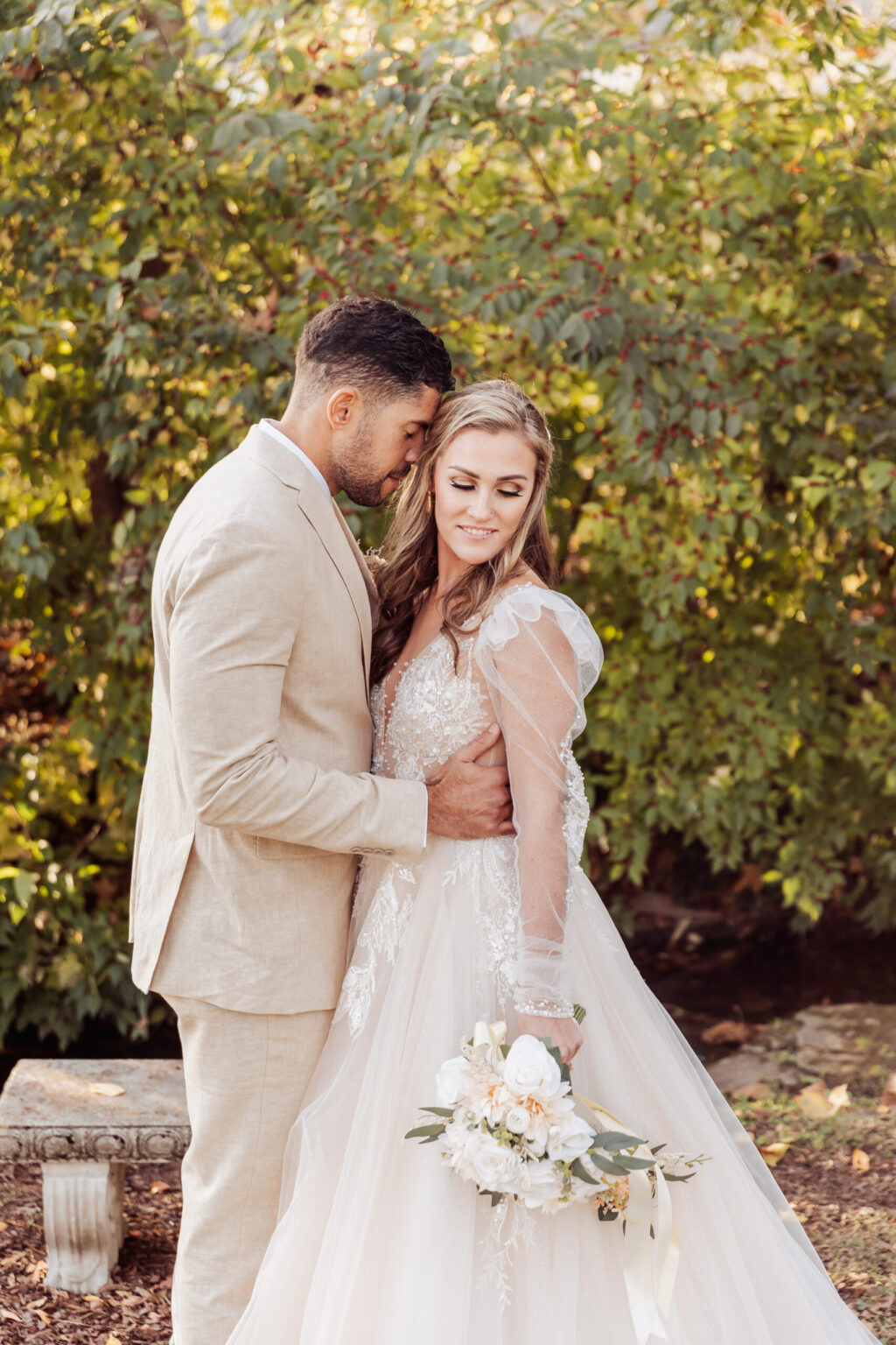 Elopement Wedding A groom wearing a light beige suit gently kisses the forehead of the bride, who is wearing an elegant, lace-adorned white wedding gown with sheer sleeves. She holds a bouquet of white roses and greenery. They stand outdoors against a backdrop of lush green foliage, capturing the romance of an intimate elopement. Elopements Inc