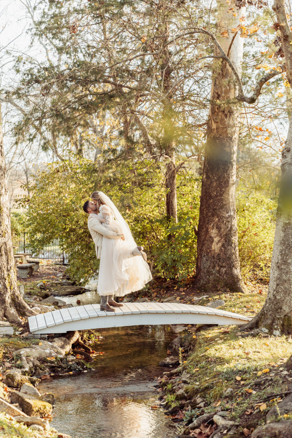 Elopement Wedding A couple dressed in wedding attire embraces on a small white bridge over a narrow creek. The groom lifts the bride, who wears a long white dress and veil. This intimate elopement is surrounded by tall trees and lush greenery. Sunlight filters through the leaves, creating a warm, romantic atmosphere. Elopements Inc