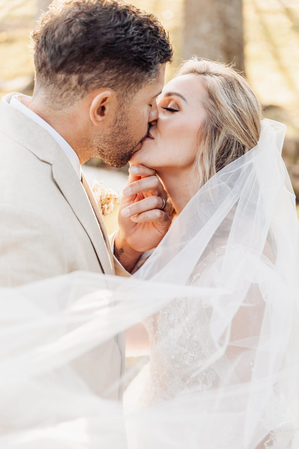 Elopement Wedding A couple shares a tender kiss in this outdoor wedding photo, part of their intimate elopement. The groom, in a light-colored suit, gently holds the bride's chin. The bride, with flowing blonde hair, wears a veil and lace dress. Sunlight filters through the trees, casting a warm glow on their special moment. Elopements Inc