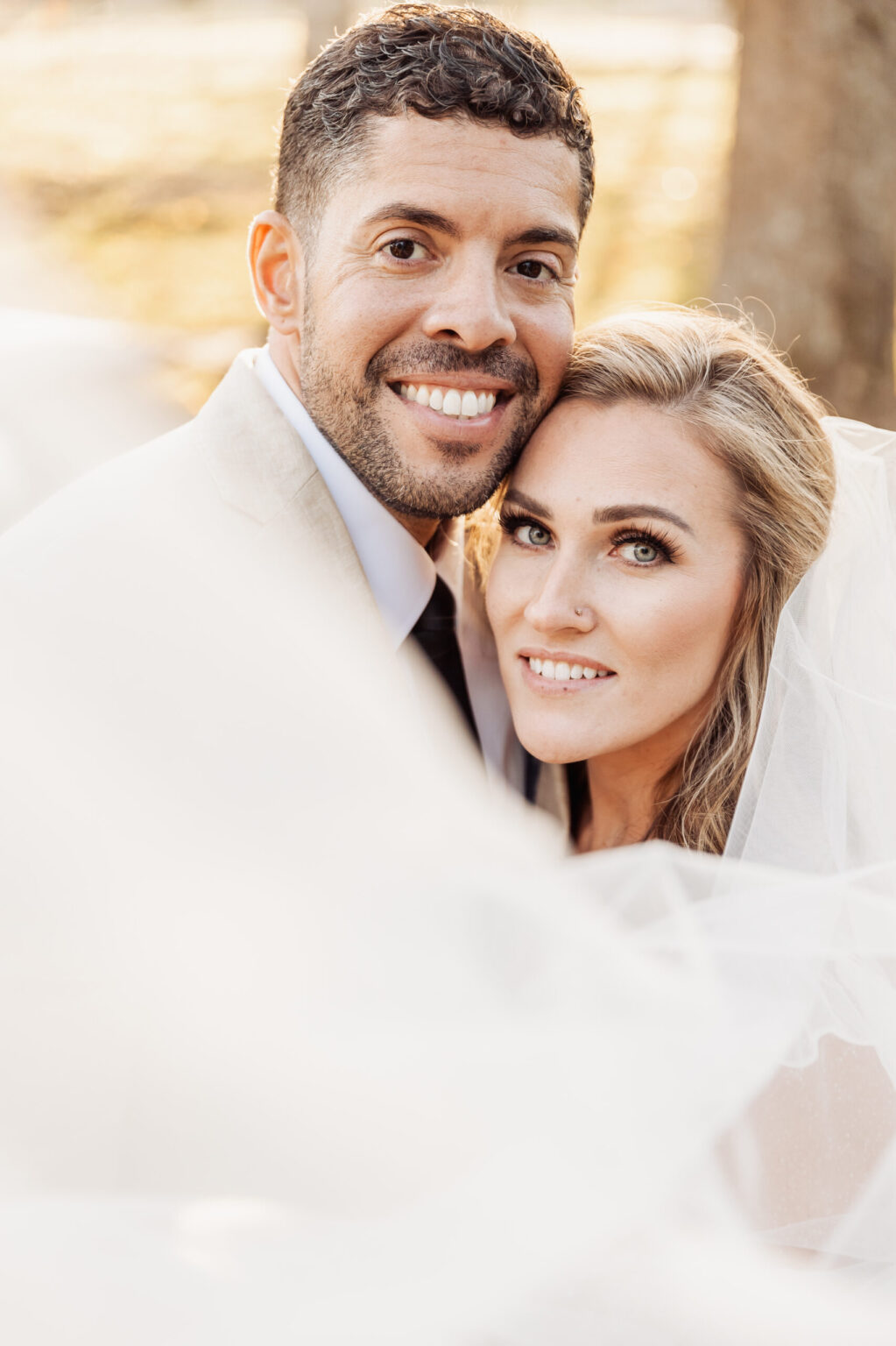 Elopement Wedding A smiling couple on their wedding day. The groom has short curly hair, a beard, and is wearing a light-colored suit with a dark tie. The bride has long, wavy blonde hair and is wearing a white dress and veil. They cuddle close after their elopement, with the veil flowing softly in the foreground and trees in the background. Elopements Inc