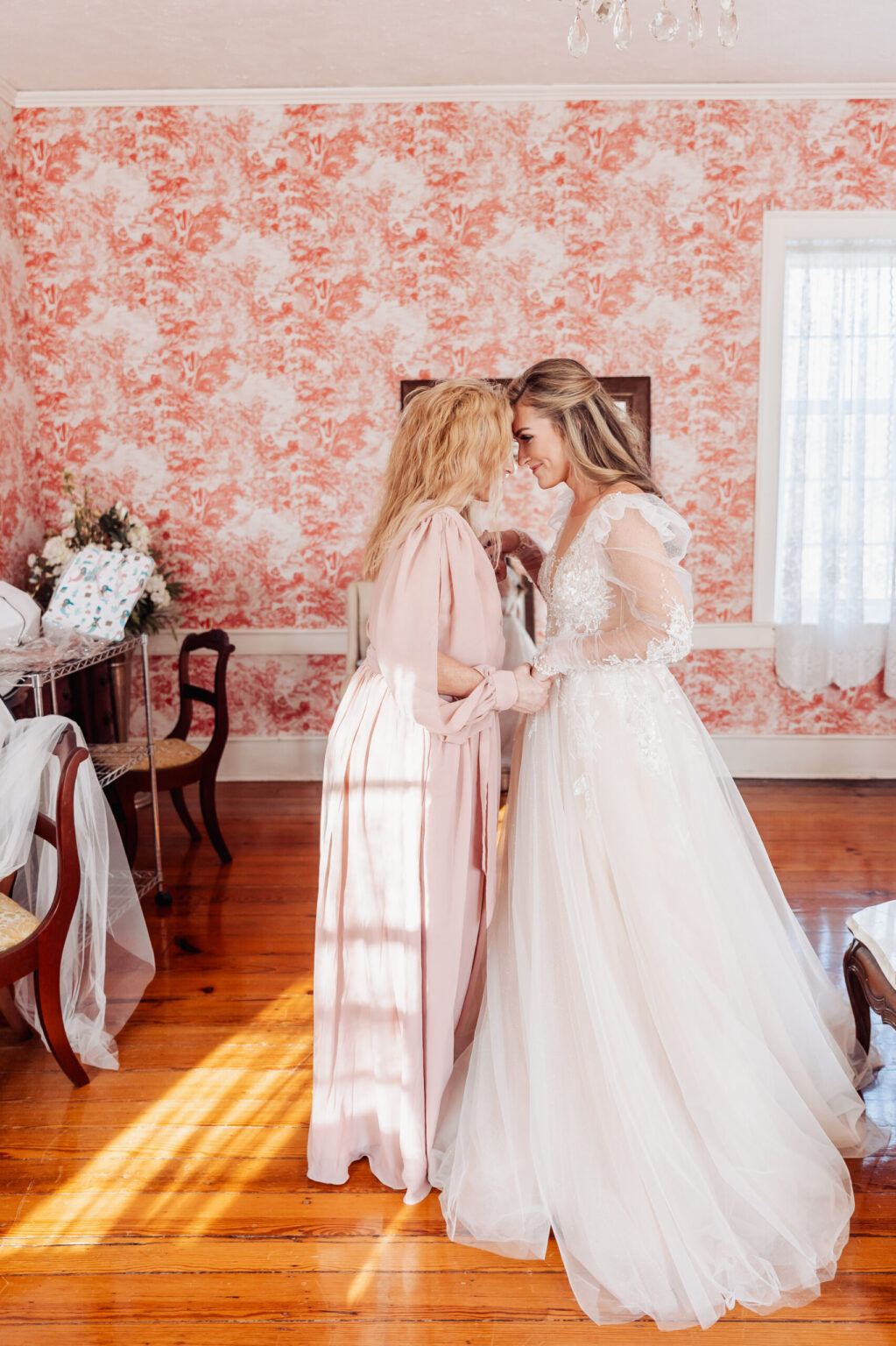 Elopement Wedding A bride in a white lace wedding gown and a woman in a light pink dress stand facing each other, holding hands and touching foreheads. The room has a bright, warm ambiance with wooden floors, floral-patterned wallpaper, and a window with white curtains allowing natural light to stream in—an intimate moment perfect for an elopement. Elopements Inc