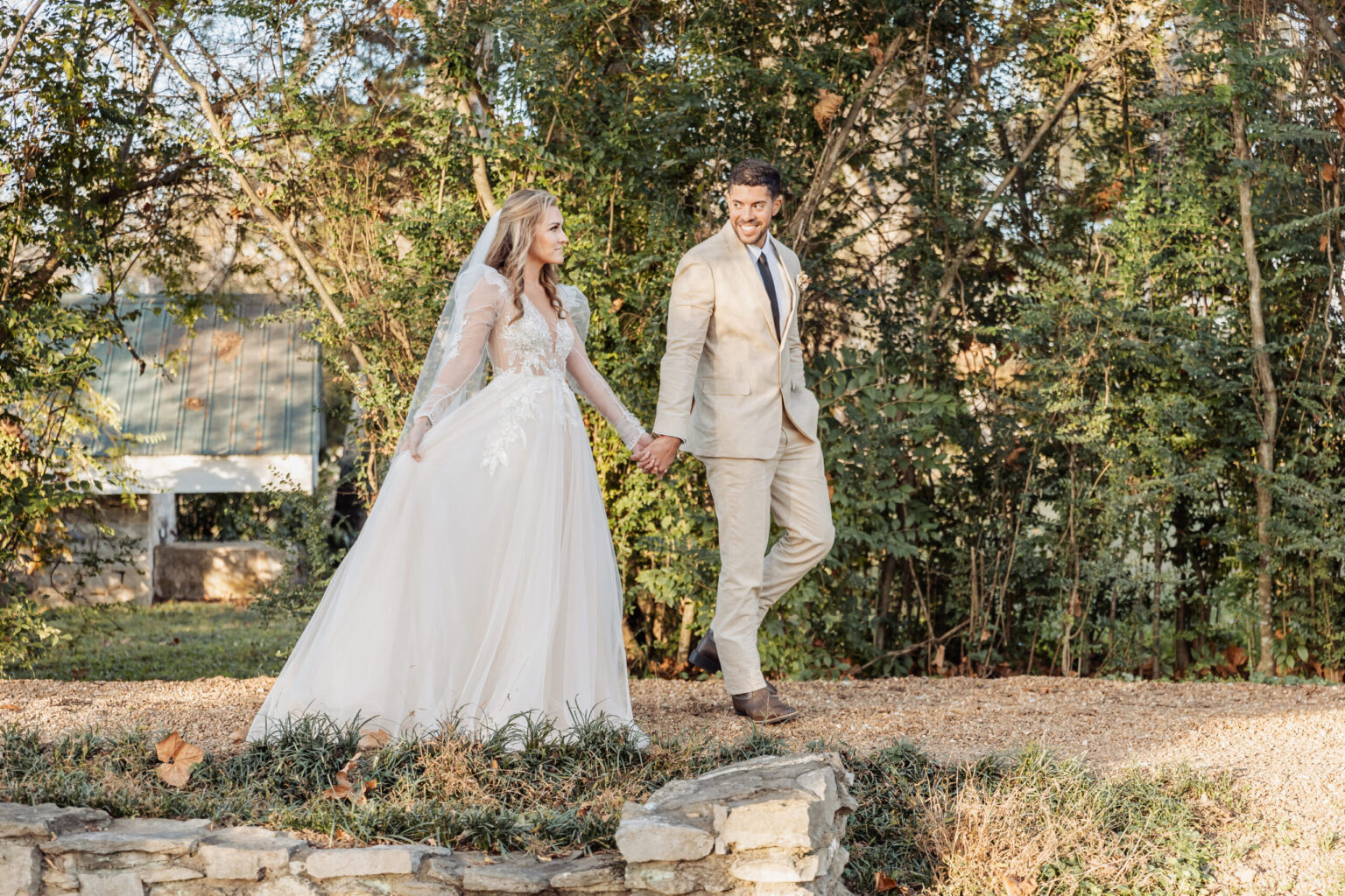 Elopement Wedding A bride in a flowing white gown with lace sleeves and a groom in a light beige suit walk hand-in-hand outdoors. They are on a stone path surrounded by lush greenery and trees with the sunlight filtering through. Their elopement is filled with joy as they smile at each other. Elopements Inc