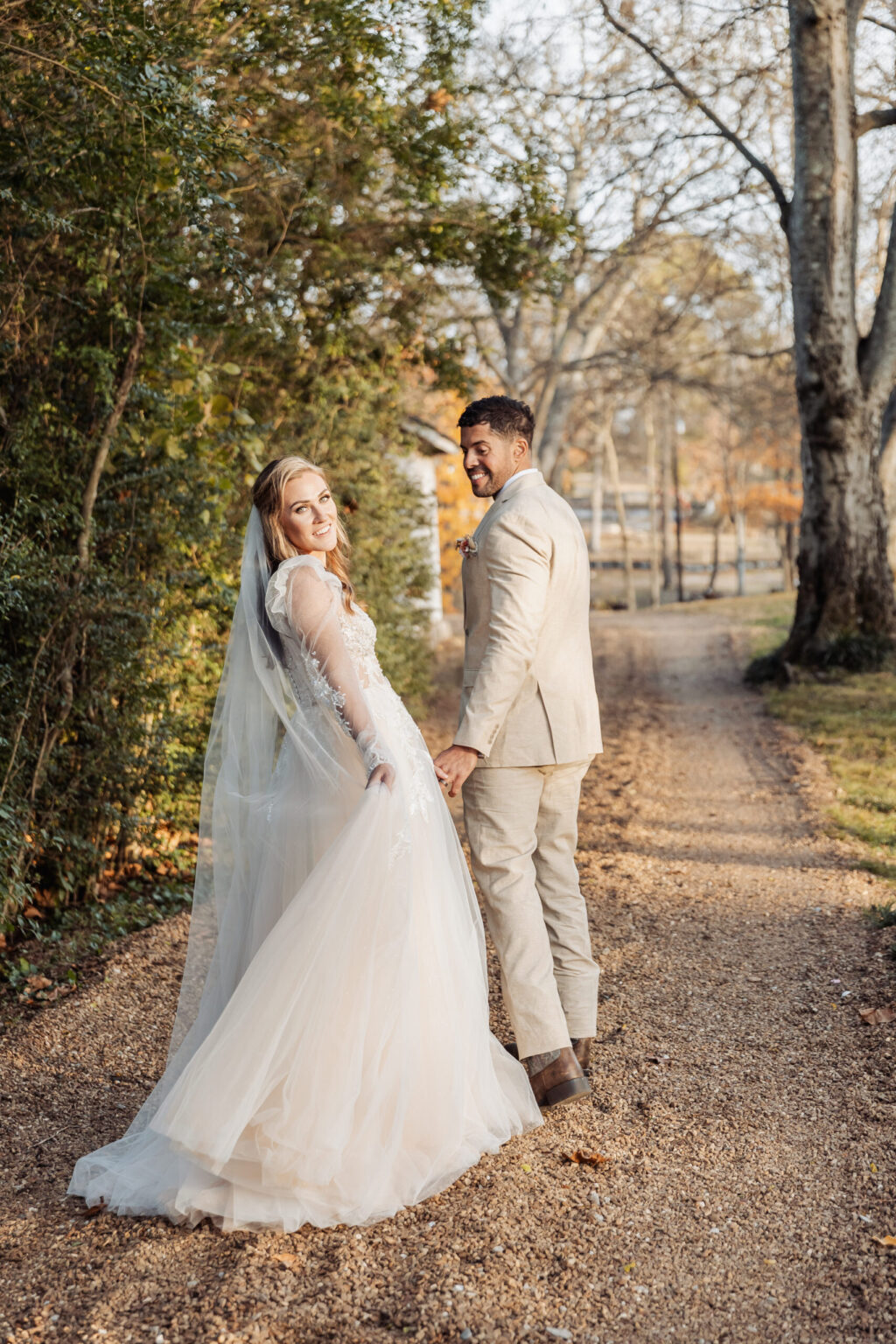 Elopement Wedding A bride and groom, having chosen to elope, walk hand-in-hand down a gravel path surrounded by trees. The bride, smiling and looking over her shoulder, wears a long white dress with a veil. The groom, in a beige suit, looks back at her. Sunlight filters through the trees, casting a soft glow on the scene. Elopements Inc