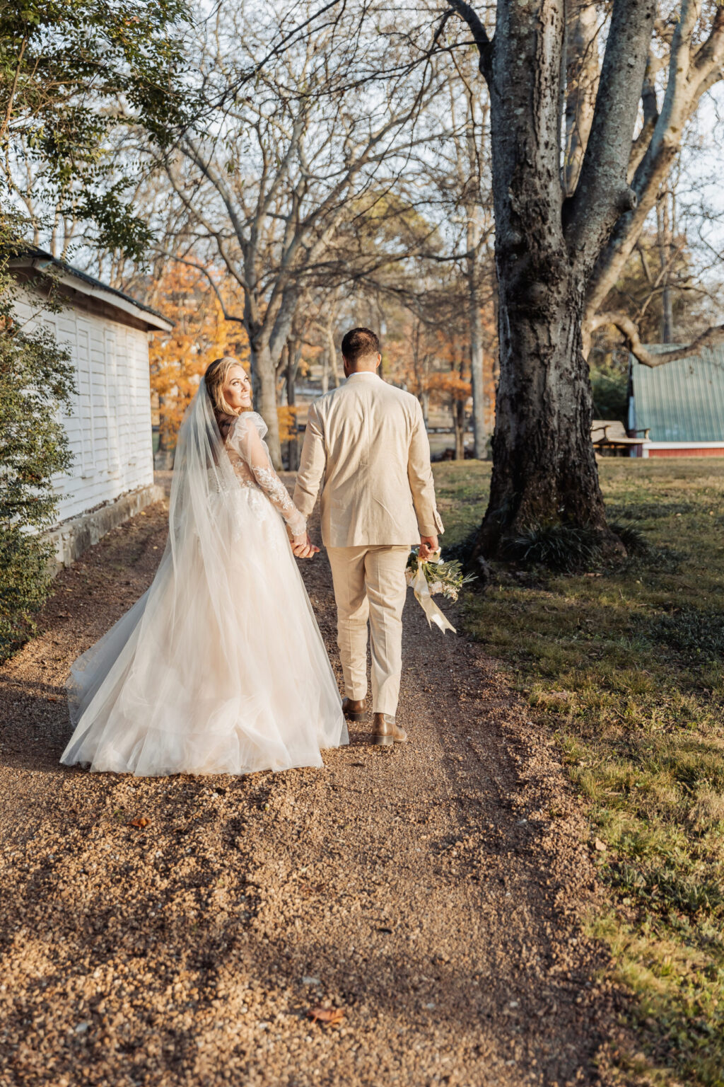 Elopement Wedding A bride and groom walk hand-in-hand on a gravel path surrounded by trees, having chosen an intimate elopement. The bride wears a flowing, white wedding dress with a long veil, while the groom is dressed in a light beige suit. It's a sunny day with autumn foliage as they stroll away from a charming white house. Elopements Inc