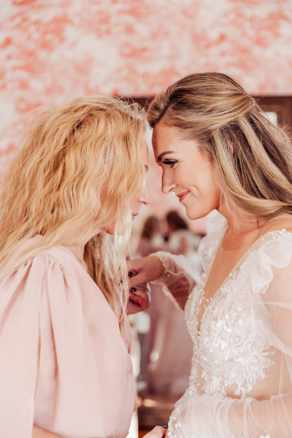 Elopement Wedding Two women are gently touching their foreheads together, sharing an intimate moment during their elopement. One woman, with long, wavy blonde hair, wears a light pink blouse, while the other, with straight, highlighted hair, wears a white floral-lace wedding dress with sheer sleeves. Both are smiling softly. Elopements Inc