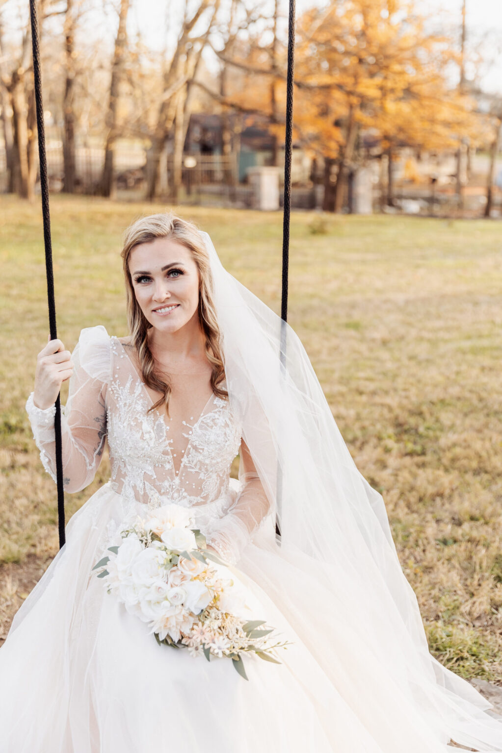 Elopement Wedding A bride in a white, floral lace wedding dress with long sleeves and a veil sits on a swing. She holds a bouquet of white and blush pink flowers. The background features an autumn landscape with bare trees and orange leaves, perfect for intimate elopements in a scenic park or garden setting. Elopements Inc