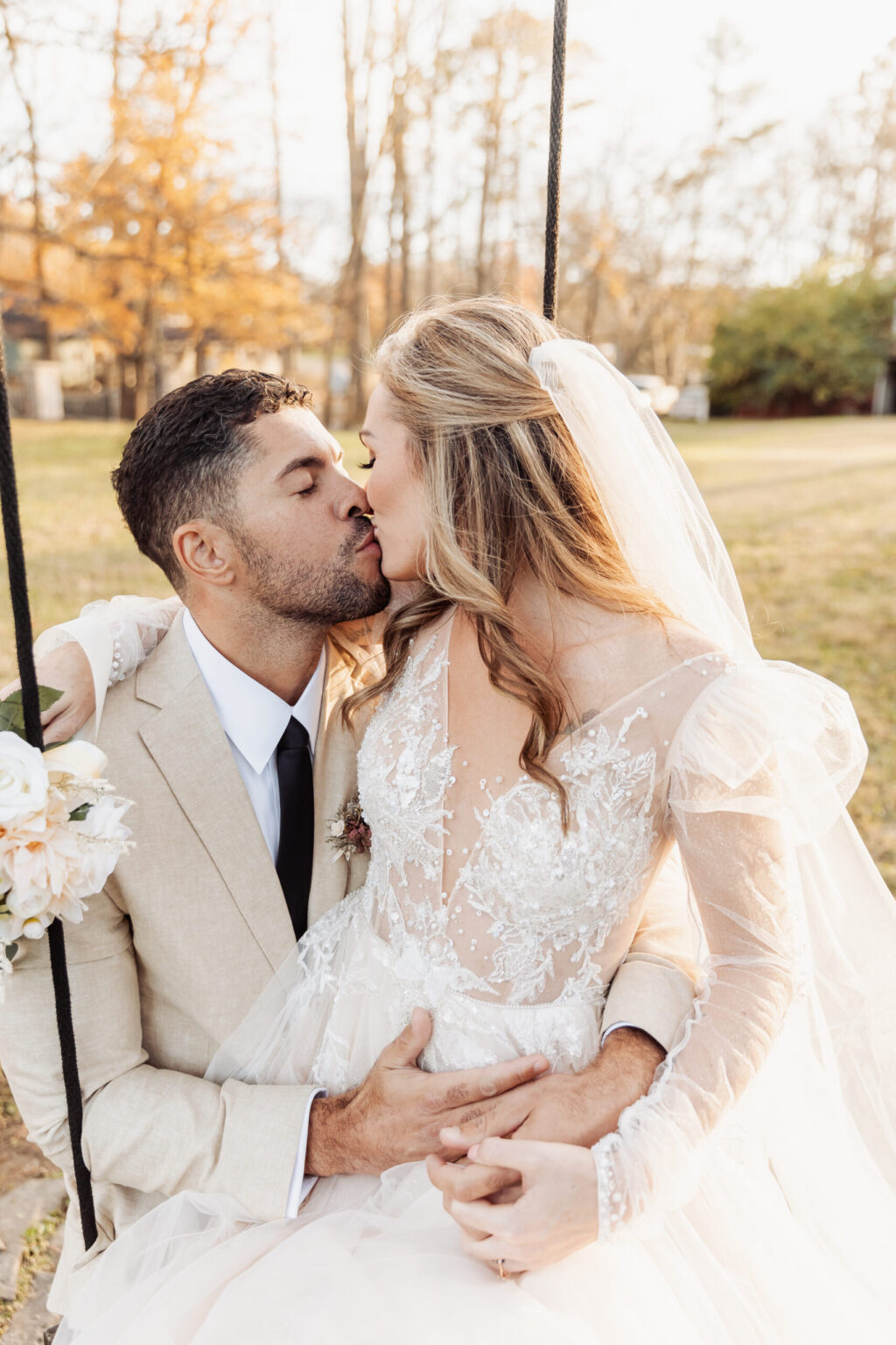 Elopement Wedding A couple shares a kiss while seated on a swing, capturing the magic of their elopement. The groom wears a light beige suit with a white shirt and black tie, and the bride is in a white lace wedding dress with sheer sleeves, holding a bouquet of flowers. They are surrounded by a grassy area with trees blurred in the background. Elopements Inc