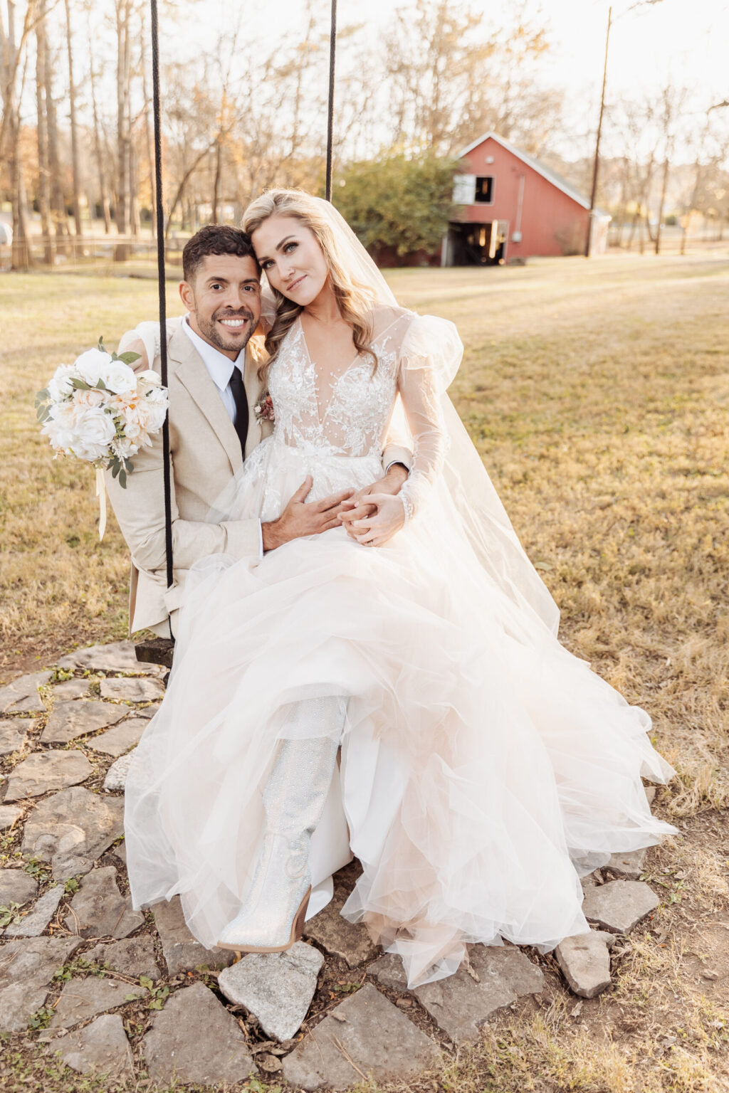 Elopement Wedding A bride and groom sit together on a wooden swing outdoors, capturing the charm of a heartfelt elopement. The groom in his beige suit holds a bouquet of white flowers, while the bride in her lacy white gown with a long veil tenderly rests her hand on her belly. An old red barn and autumn trees frame their intimate moment. Elopements Inc