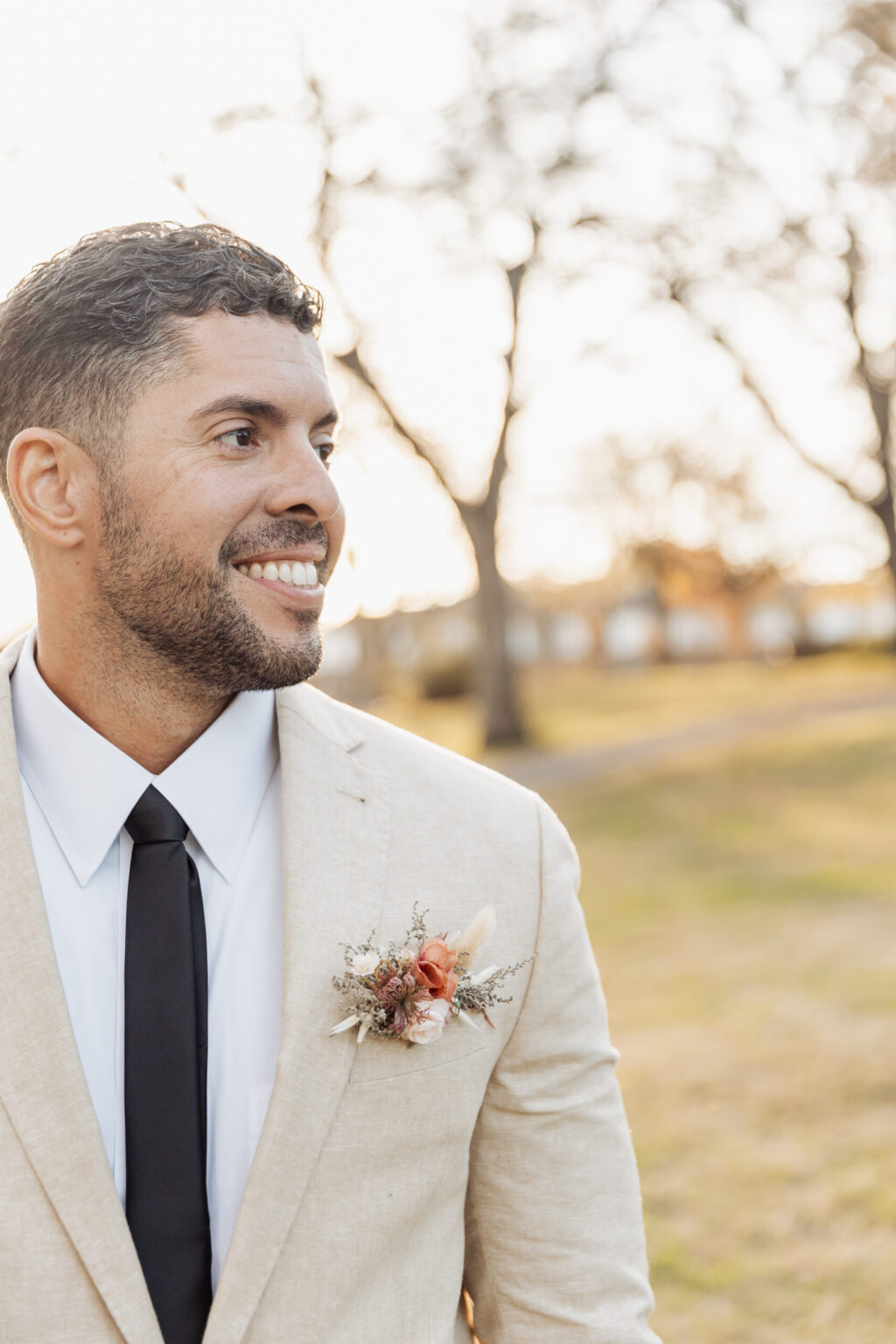 Elopement Wedding A man with short dark hair and a beard is smiling and looking to his left. He is wearing a light beige suit jacket, white dress shirt, and a black tie. A small boutonniere with red, white, and greenery adorns his lapel. The grassy area and sparse, bare trees in soft daylight set the perfect scene for an elopement. Elopements Inc