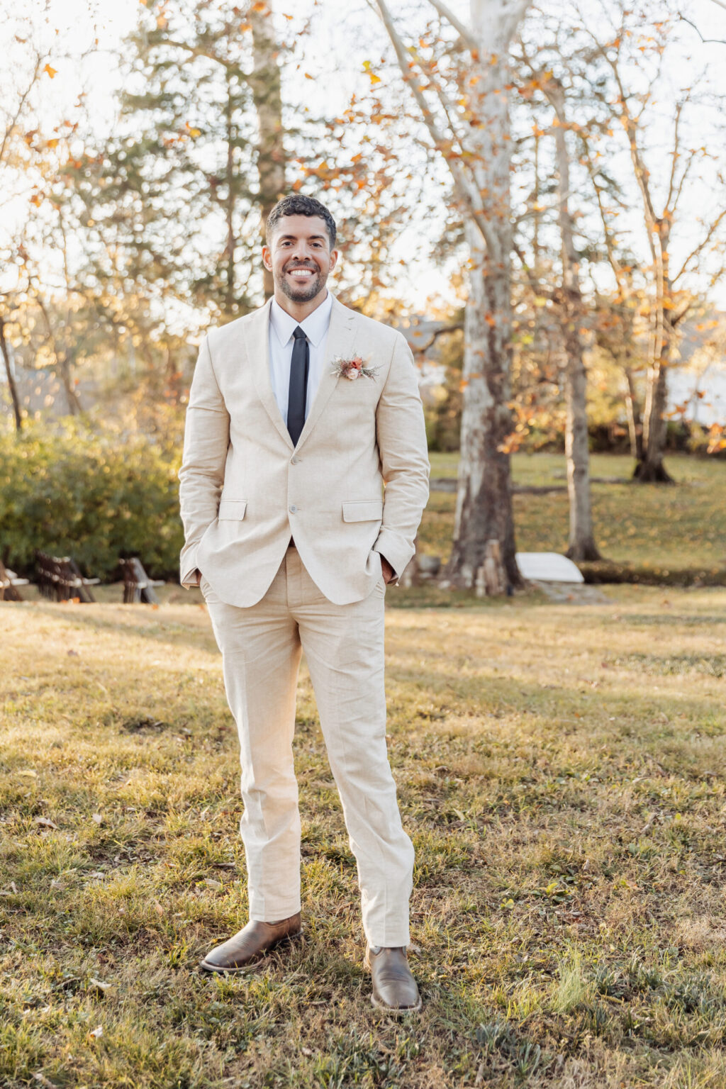 Elopement Wedding A man with short hair and a beard is standing outdoors on grassy ground with trees and a sunlit background. He is wearing a beige suit with a white shirt, a black tie, and brown shoes. A small floral boutonniere is pinned to his left lapel. He is smiling and has his hands in his pockets, ready for an intimate elopement. Elopements Inc