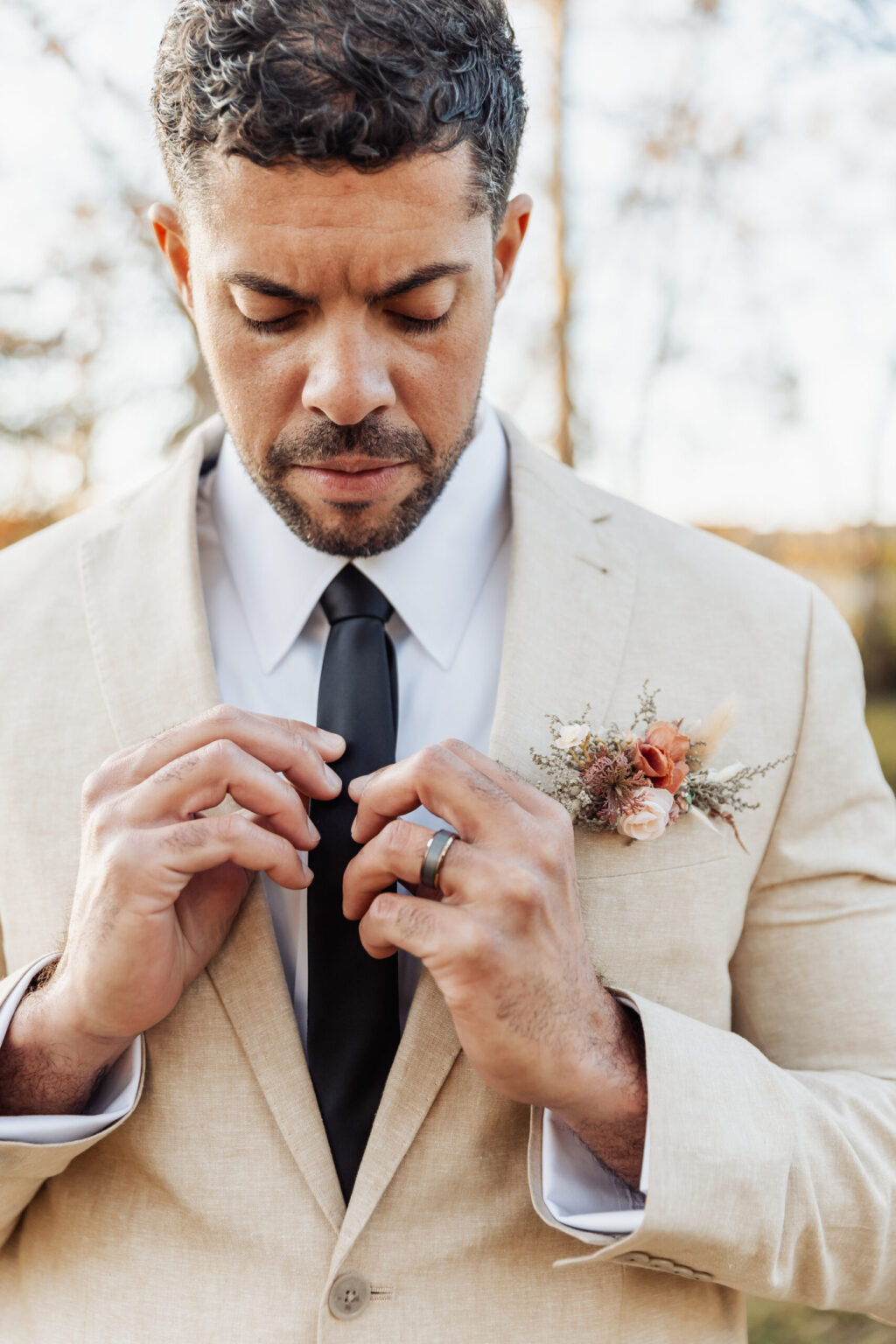 Elopement Wedding A man with short, curly hair and a beard adjusts his black tie while wearing a beige suit jacket and a white dress shirt. A small boutonniere with pink and beige flowers is pinned to his jacket, hinting at an intimate elopement. The background is blurred with an outdoor setting, hinting at trees and natural light. Elopements Inc