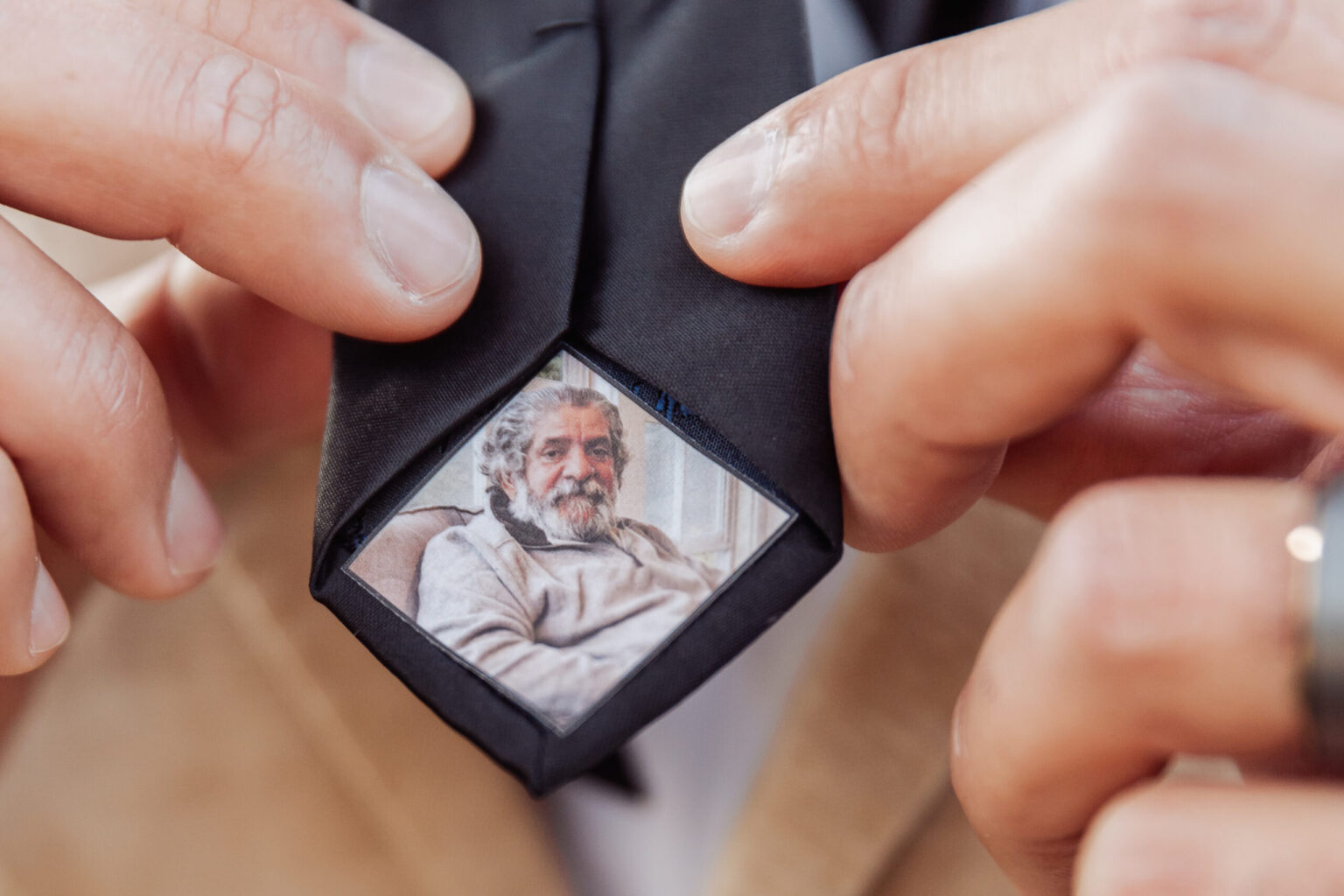 Elopement Wedding A person holding the end of a black necktie, revealing a small photo of an older man with grey hair and a beard, dressed in a beige sweater. The older man, possibly reminiscing about his youthful elopement, is sitting in a casual, relaxed pose. The person holding the tie is wearing a beige jacket. Elopements Inc